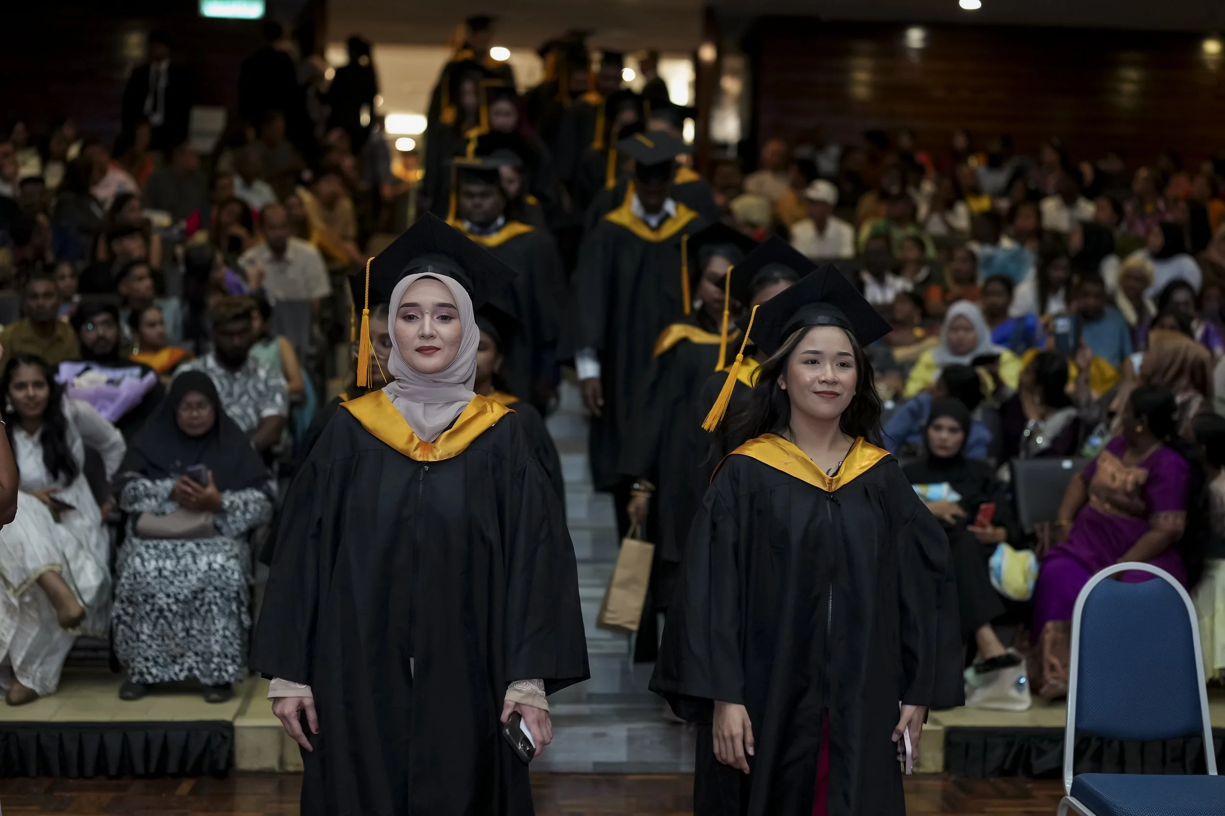 University graduation ceremony with students in caps and gowns walking across stage and audience watching.