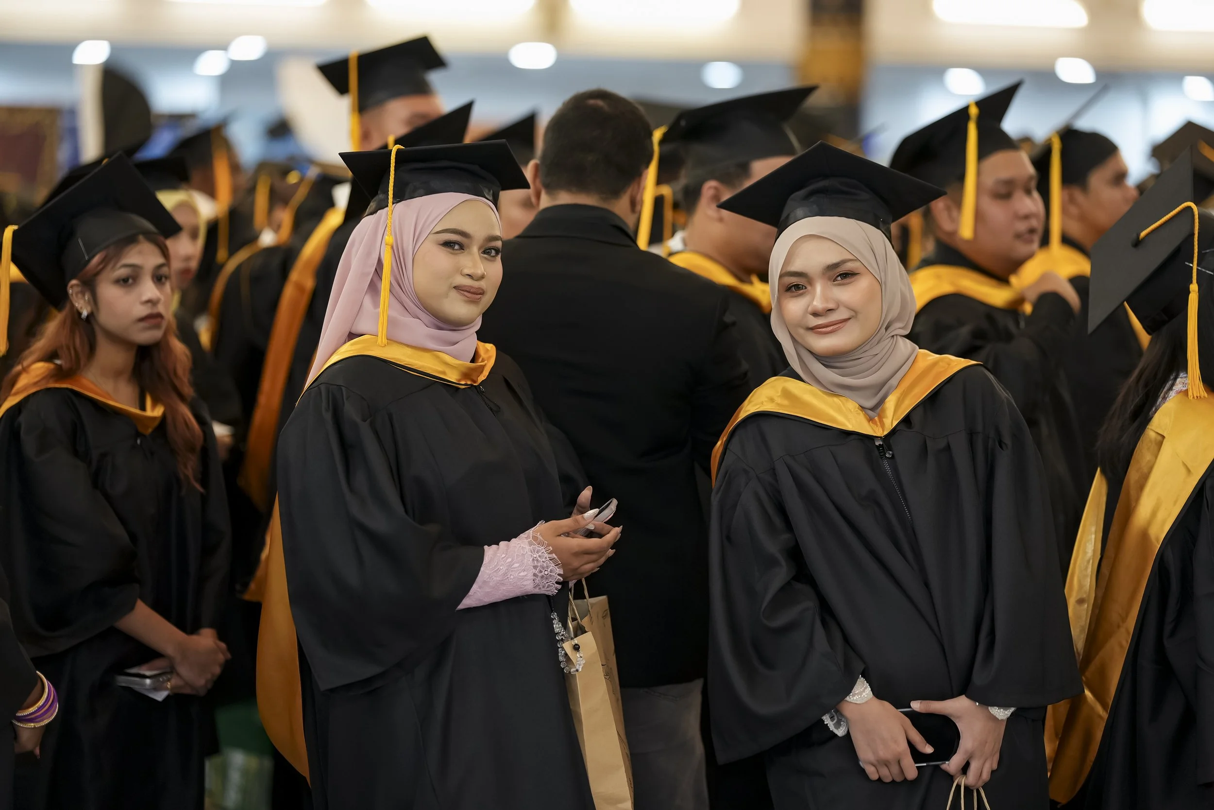 Group of diverse graduates wearing black caps and gowns, some with yellow sashes, at a graduation ceremony.