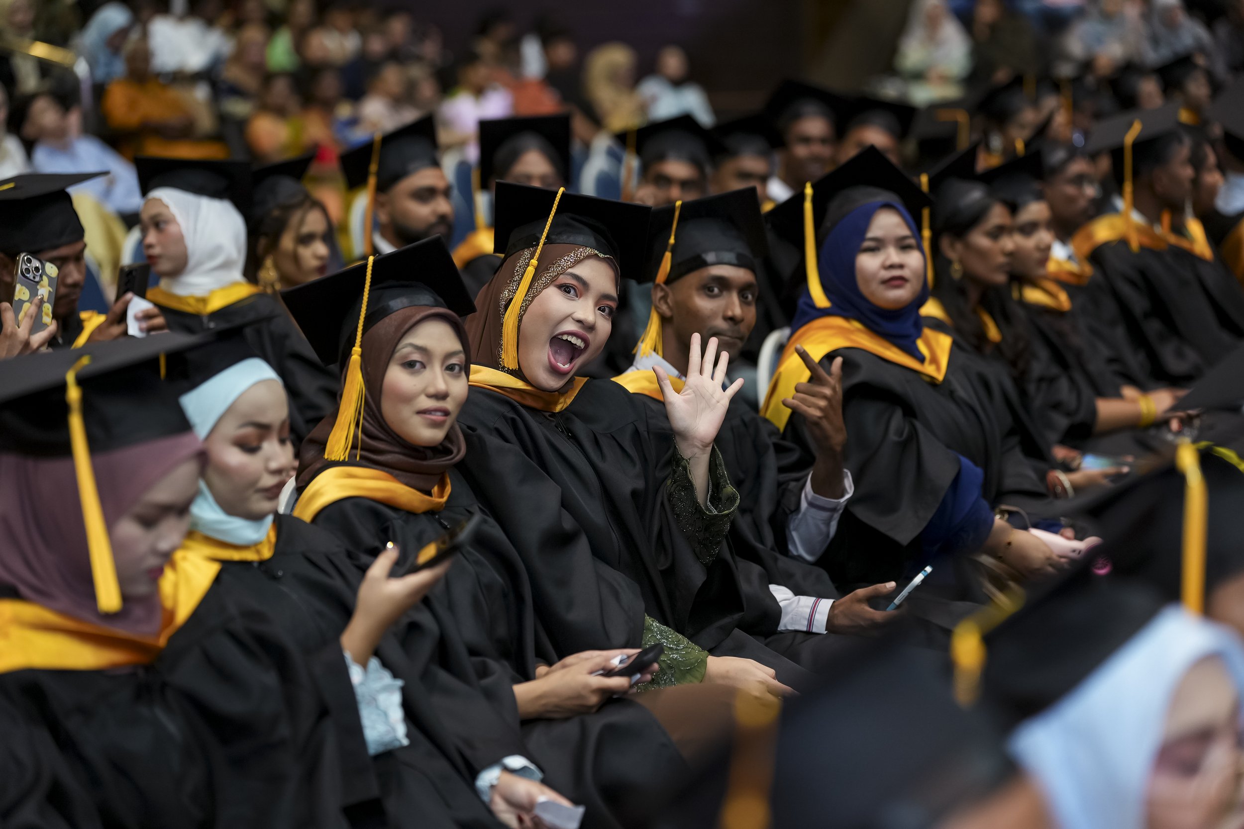 Group of graduates seated during a graduation ceremony, wearing black caps and gowns with yellow trim, some women wearing headscarves, one woman smiling and waving.