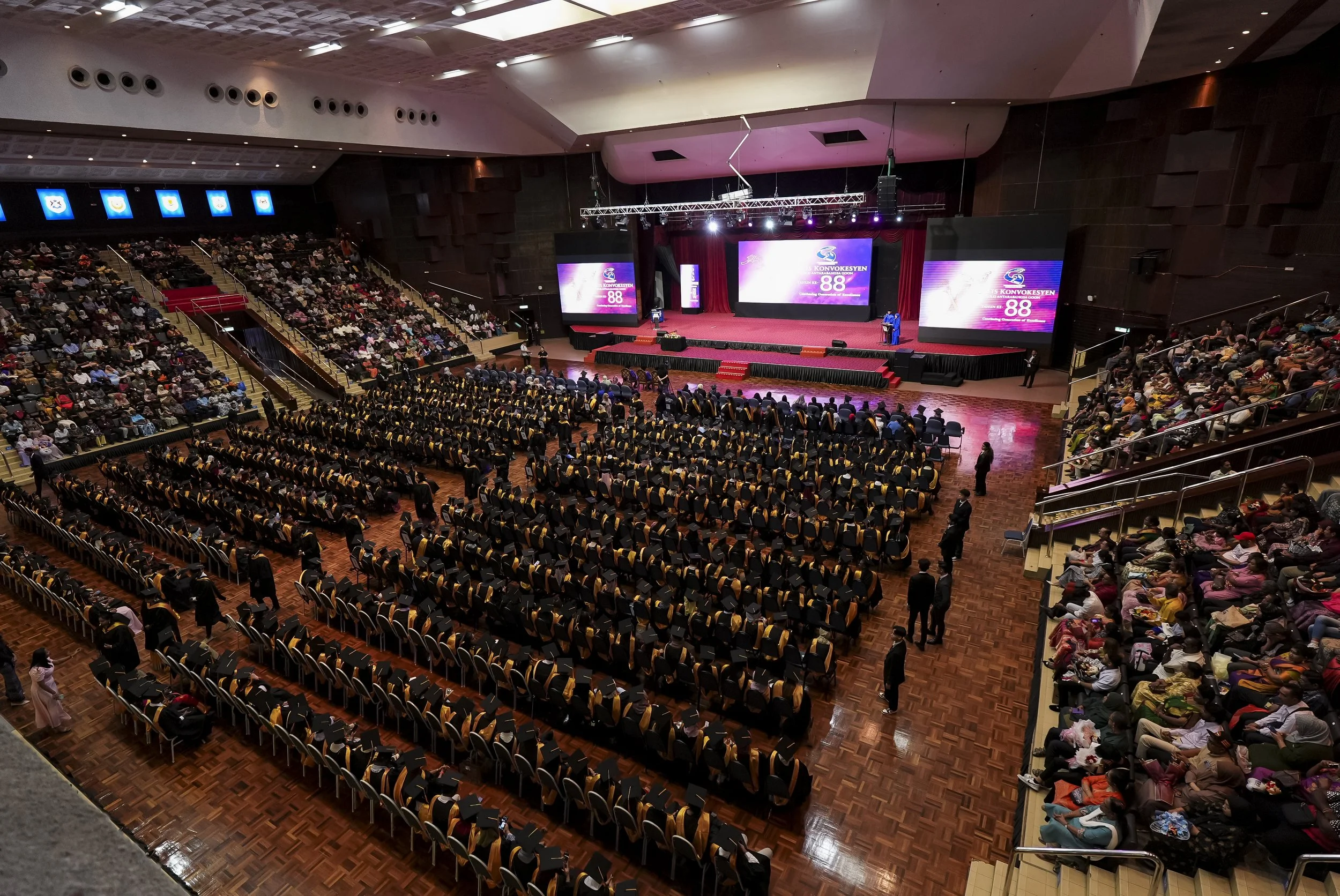 A large indoor auditorium filled with seated attendees, a stage at the front with speakers and large screens displaying the event's logo, which reads 'KONVOKESYEN' and '88'. People are dressed formally, and there are bright lights illuminating the st
