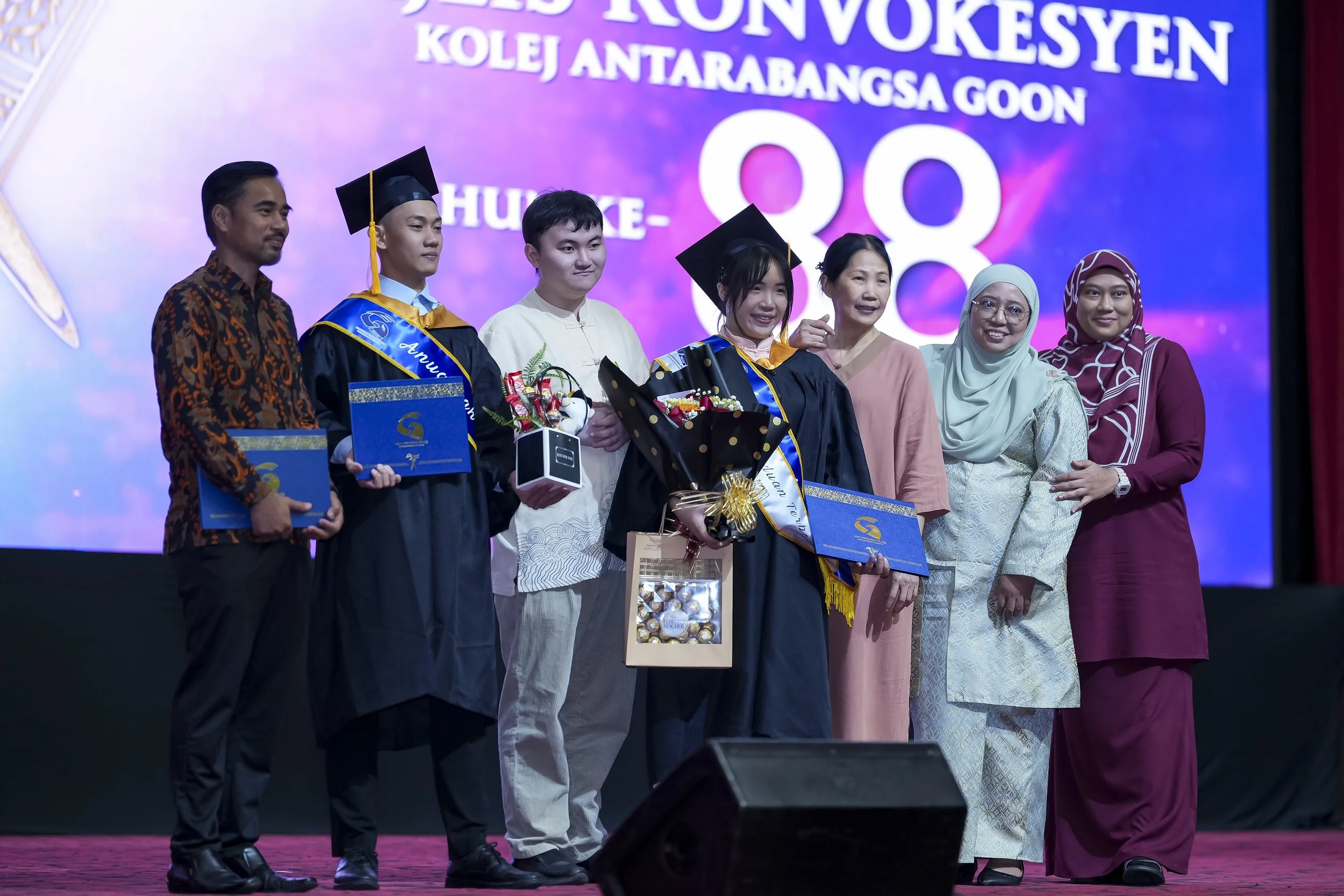 A group of eight people, including graduating students in caps and gowns, standing on stage during a graduation ceremony. The background displays text in Indonesian related to the event.