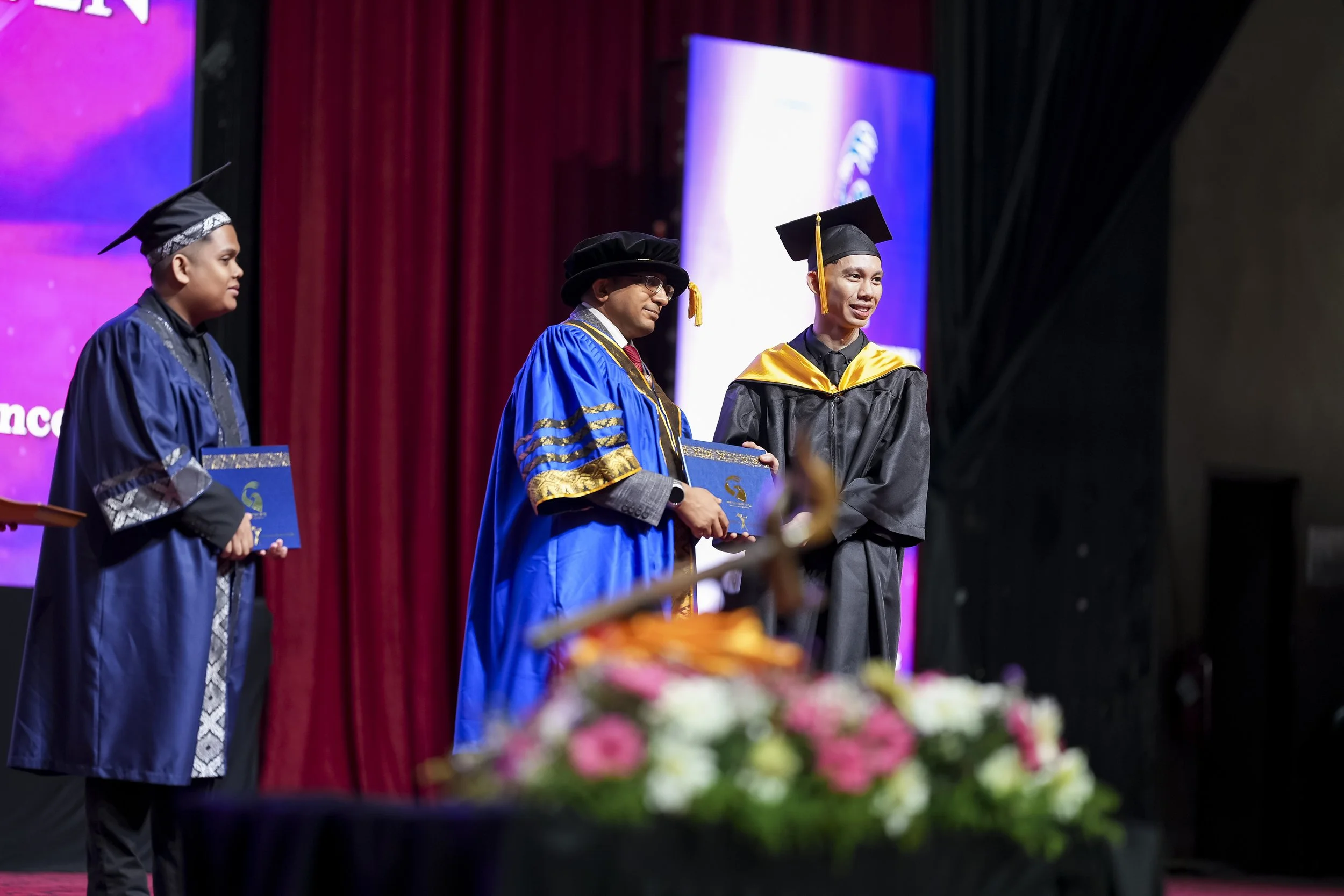 A graduation ceremony where a young man in a black cap and gown is receiving a diploma from a man in academic regalia. Other graduates in caps and gowns are standing nearby, and colorful flowers are in the foreground.