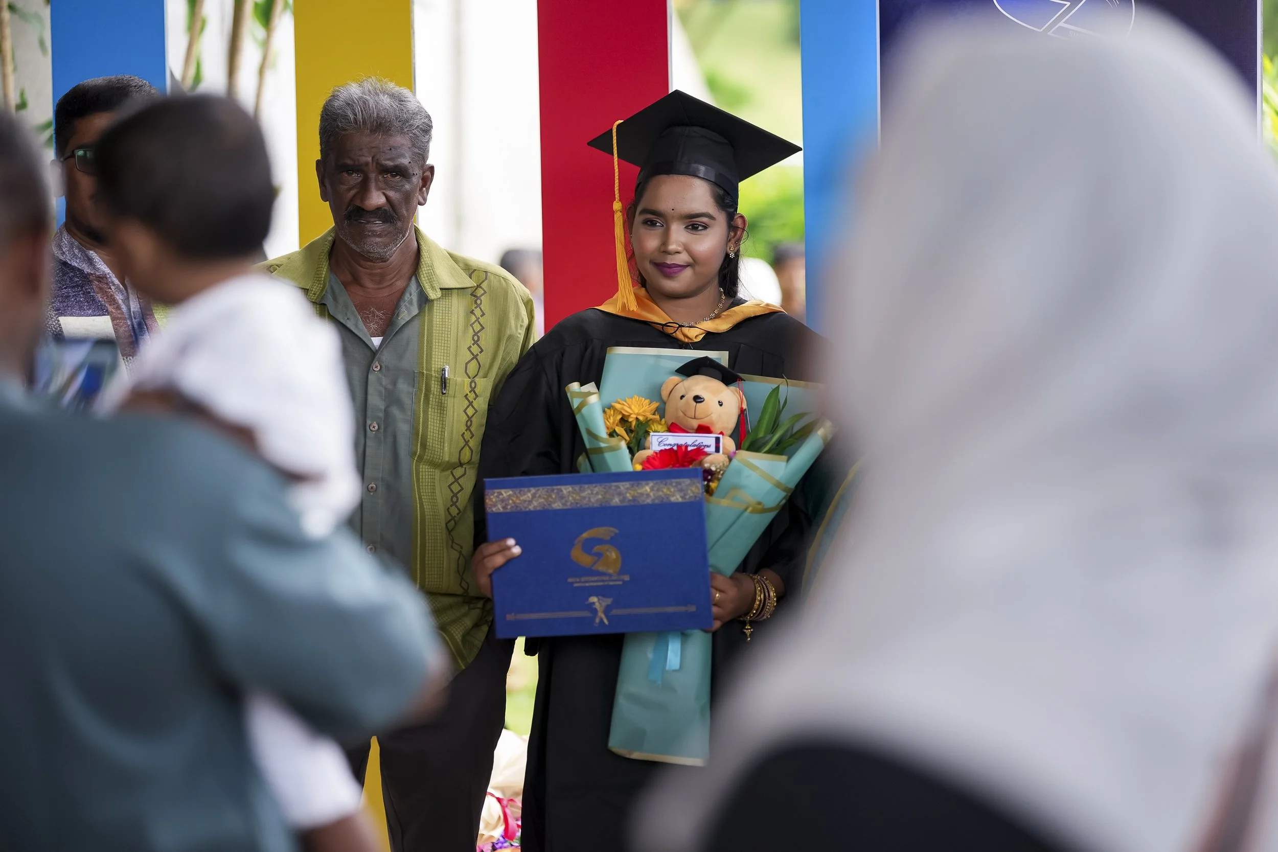 Young woman in graduation cap and gown holding flowers and a teddy bear, standing next to an older man at an outdoor graduation ceremony.