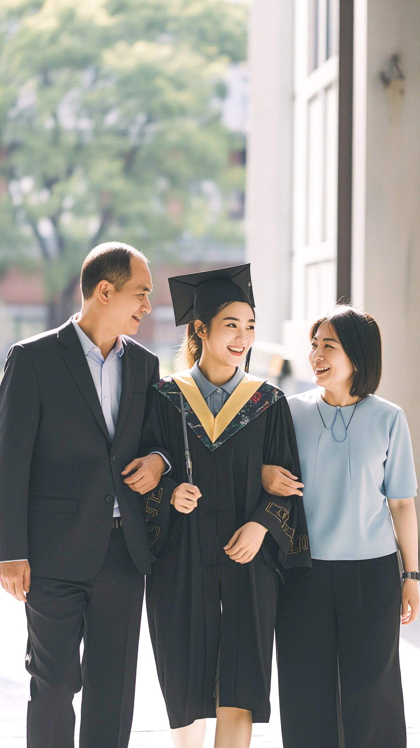 A young woman in a graduation cap and gown, smiling and walking arm-in-arm with an older man and woman, both dressed in formal attire. They are in a well-lit environment with large windows and trees outside.