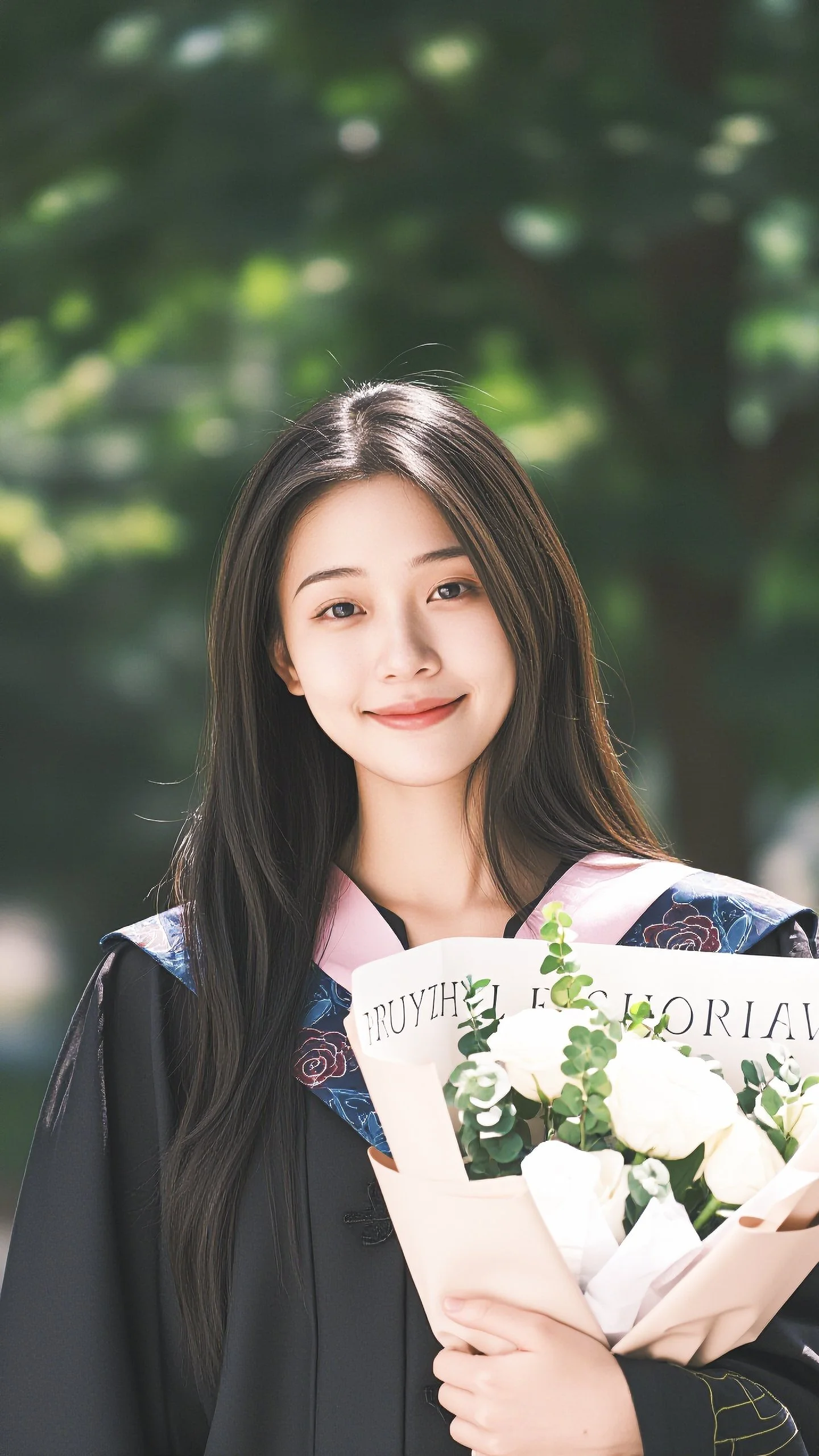 A young woman with long dark hair and a light complexion, smiling, wearing a graduation gown and holding a bouquet of white flowers wrapped in pink paper, outdoors with blurred green foliage in the background.