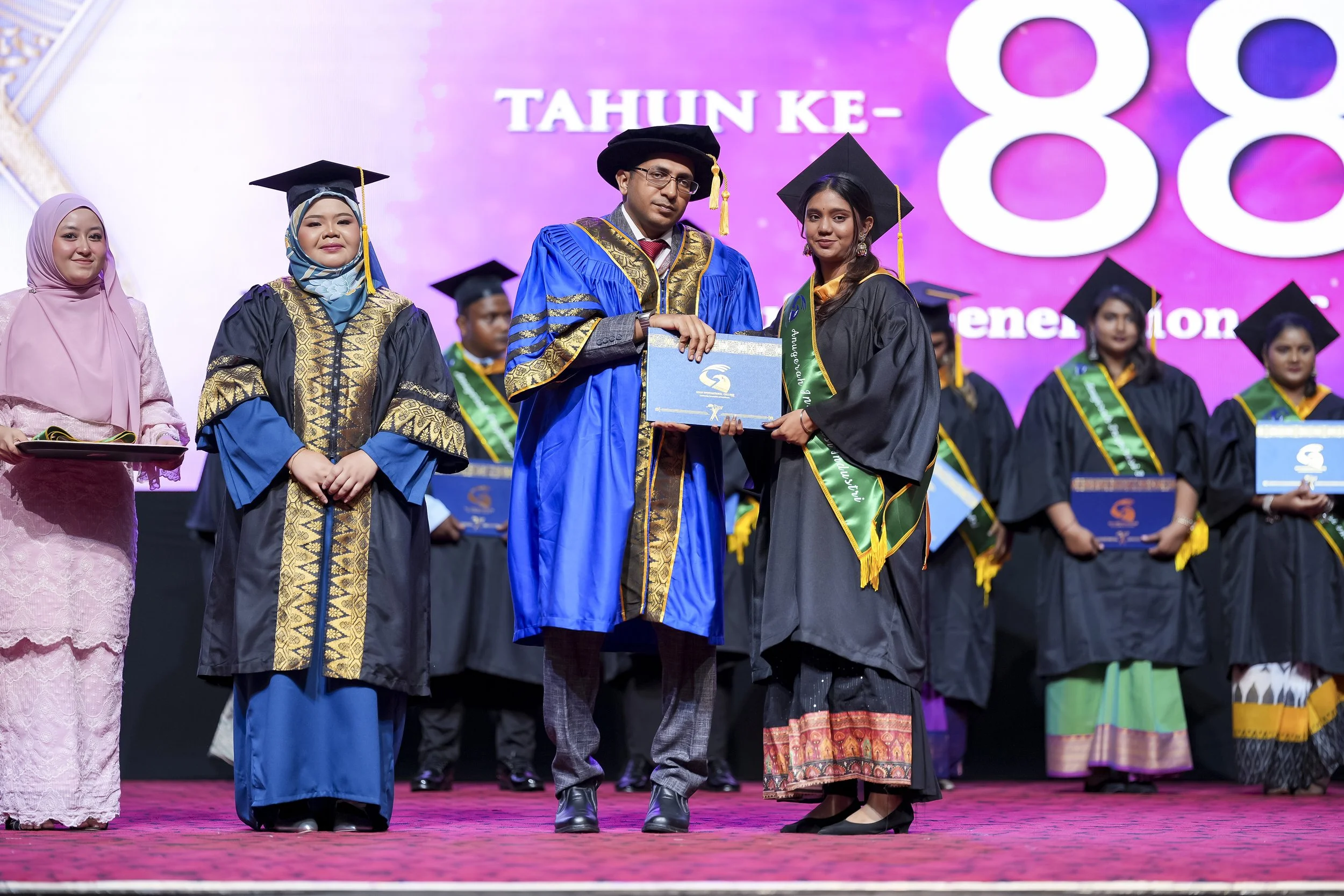 Group of graduates in black and blue caps and gowns receiving a diploma on stage during graduation ceremony.