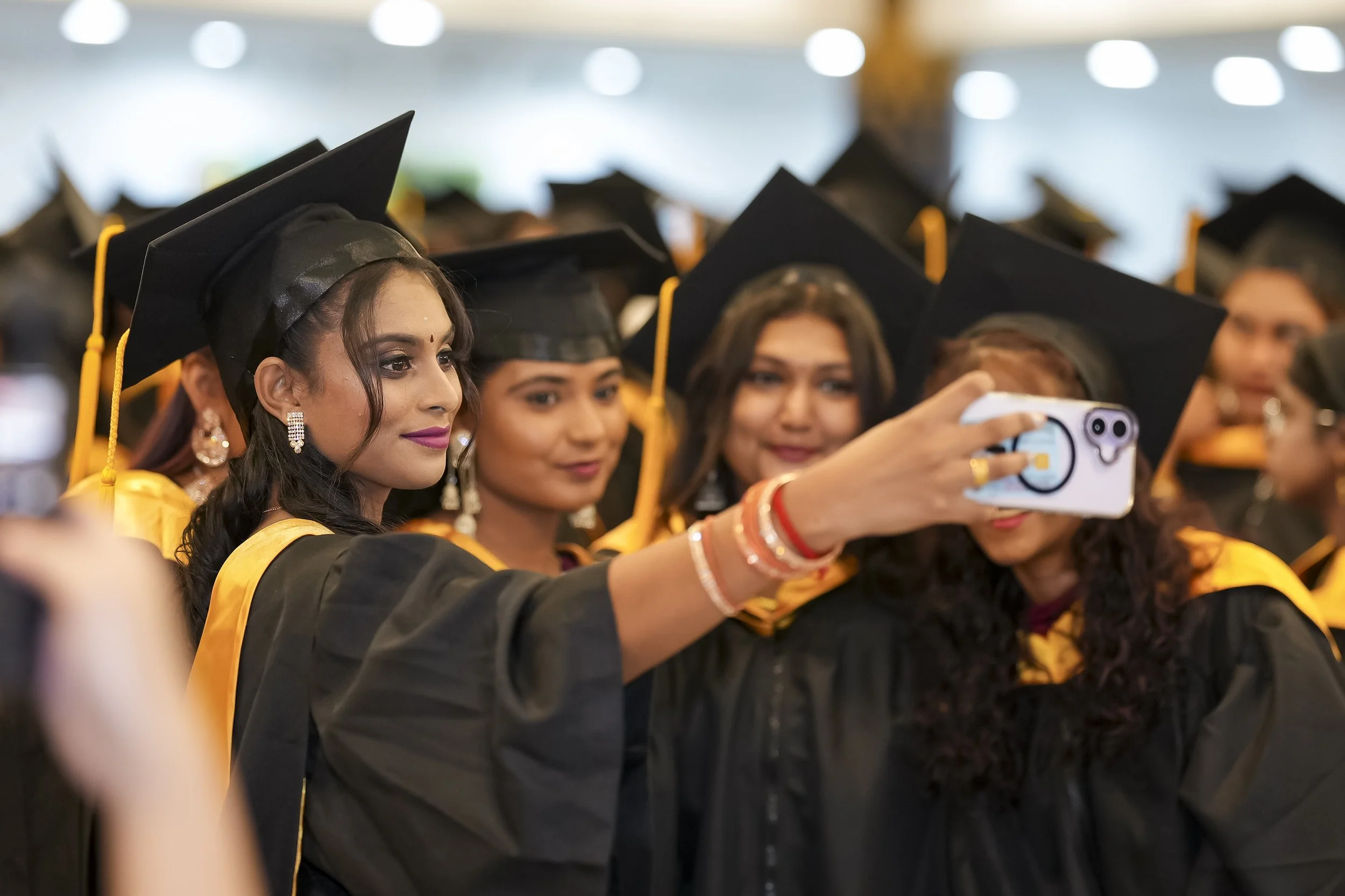 Group of diverse graduates wearing black caps and gowns, taking a selfie at graduation ceremony.