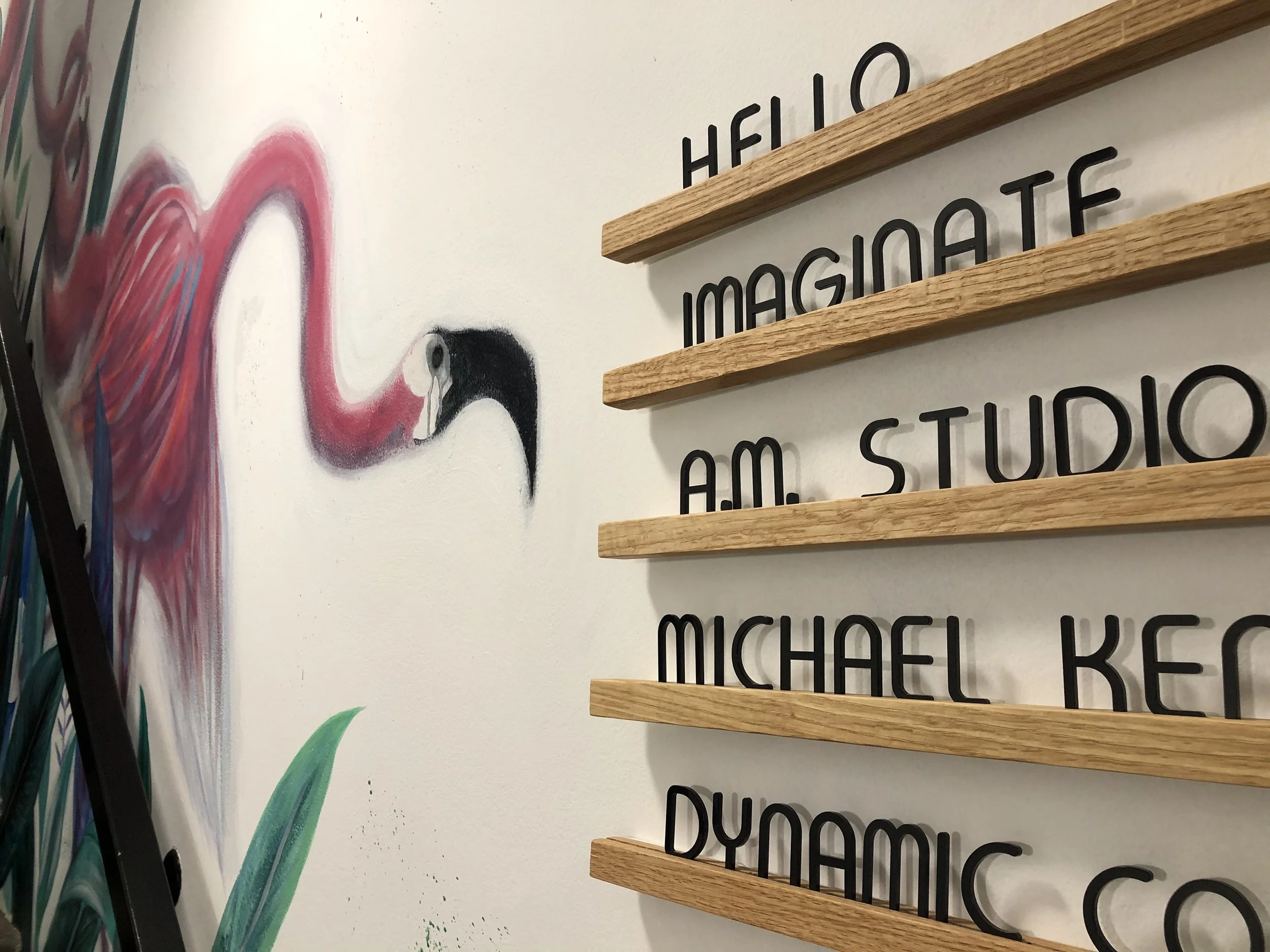 Wall art of a flamingo with pink and white feathers near a painted green leaf, next to wooden letter board with black text reading 'Hello'