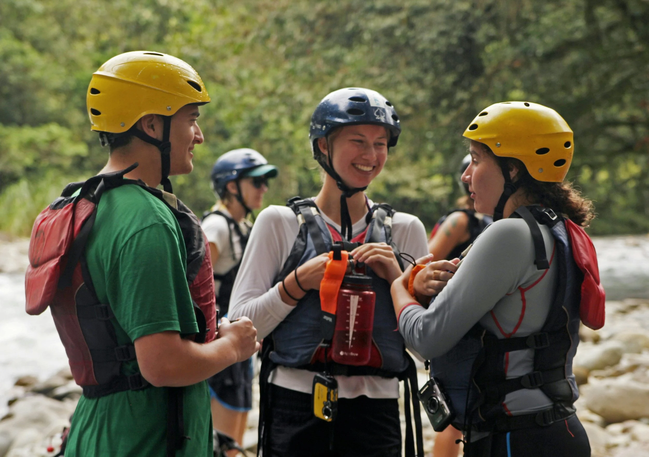 Group of people wearing helmets and life jackets, preparing for outdoor adventure near a river.