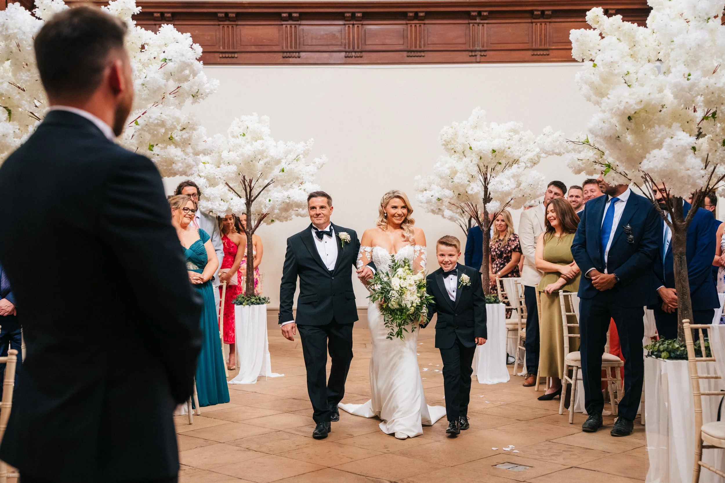 Bride walking down the aisle with her dad and son in the Main Hall at Wollaton Hall in Nottingham