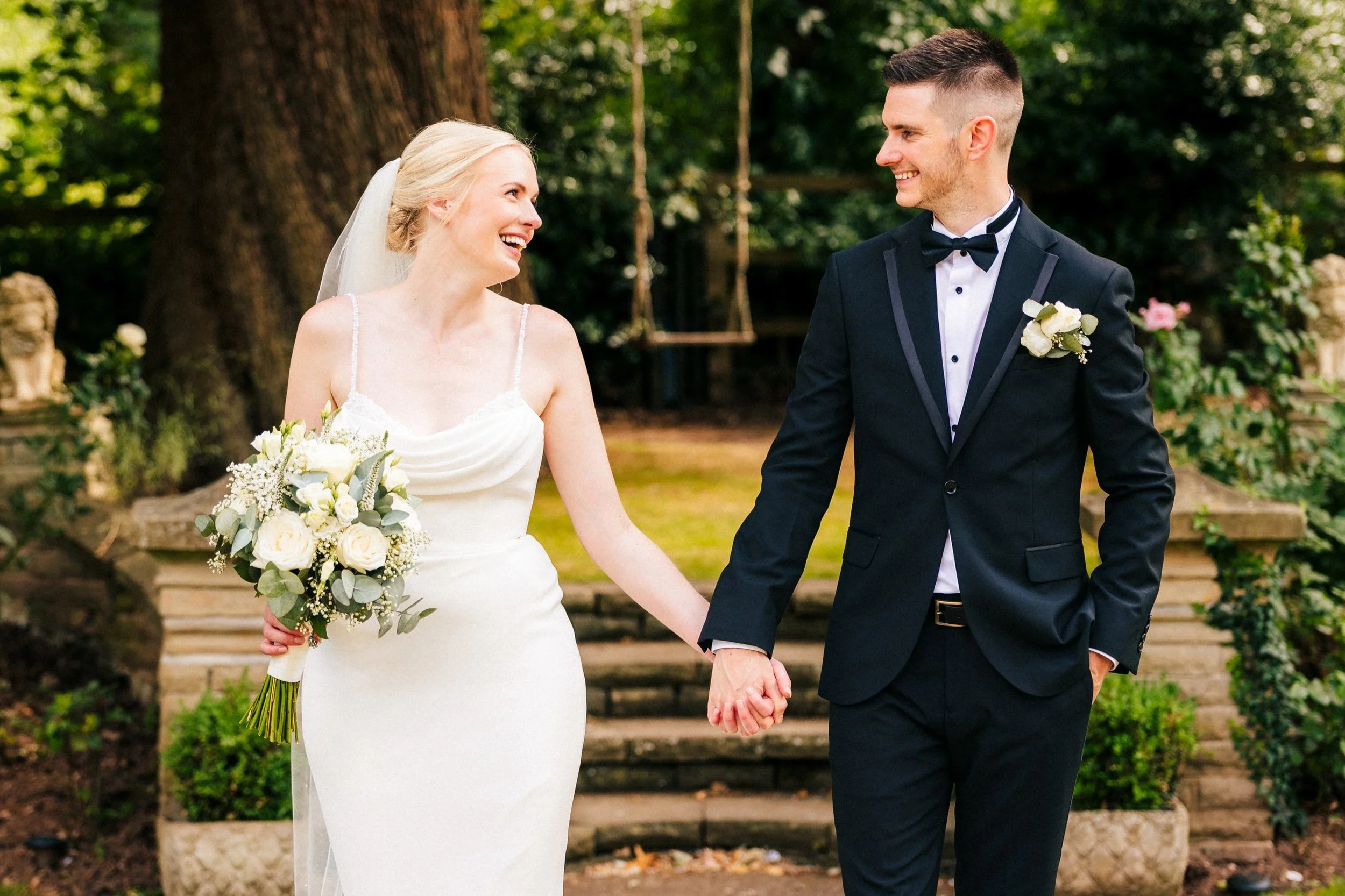 Bride and groom walking hand in hand and smiling at each other in the gardens of Woodborough Hall, captured by Andy Saywell — Nottingham wedding photographer at www.saywellhq.co.uk.