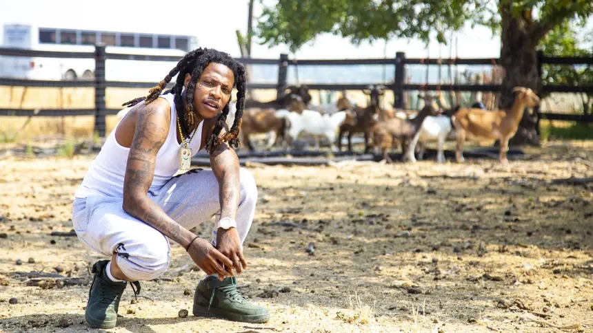 A man with dreadlocks, tattoos, and wearing a white sleeveless shirt, white pants, and green boots crouches on the dirt ground of a farm. Behind him, there are goats and a wooden fence, with trees in the background.