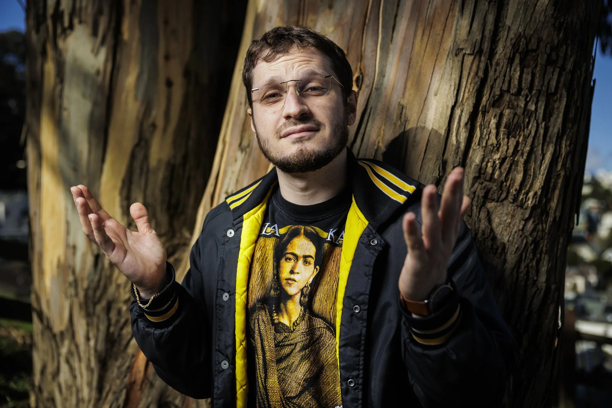 A man with glasses and a beard standing outdoors against a large tree trunk, wearing a black jacket with yellow accents and a graphic t-shirt featuring a woman.