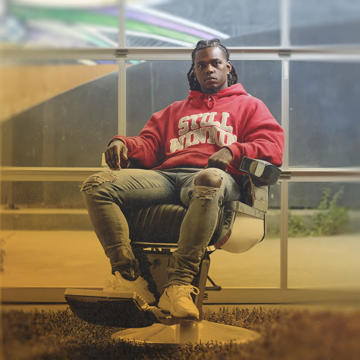 Person sitting in a chair wearing a red hoodie with 'Still Winnin' text, ripped jeans, and white sneakers. Modern interior with large windows in the background.
