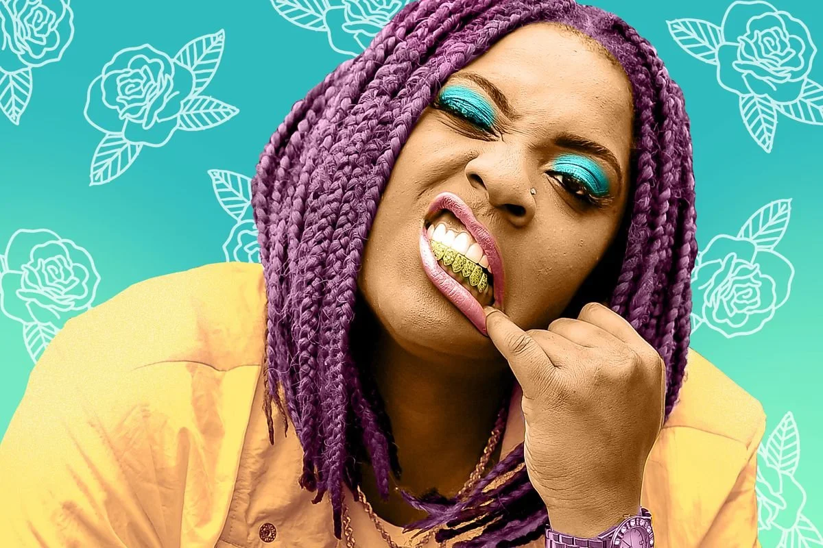 Kamaiyah with purple braids showing a gold grill, wearing bright turquoise eyeshadow, in front of a floral patterned turquoise background.