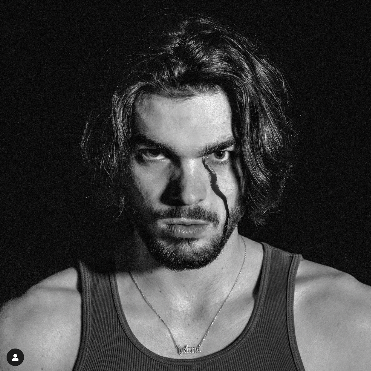 Black and white portrait of a man with long hair and a beard, wearing a tank top and necklace, with a striking shadow across his face.