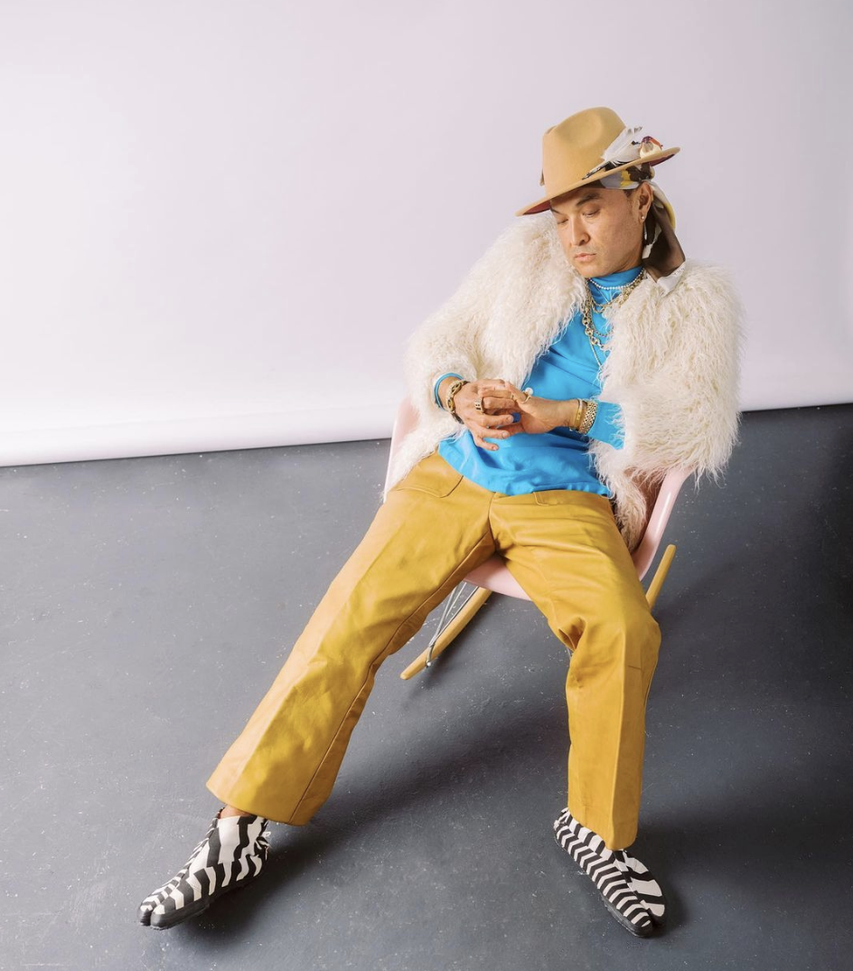Lyrics Born wearing a hat with feathers, furry jacket, blue shirt, yellow pants, and zebra-patterned shoes, sitting on a chair in a studio setting.