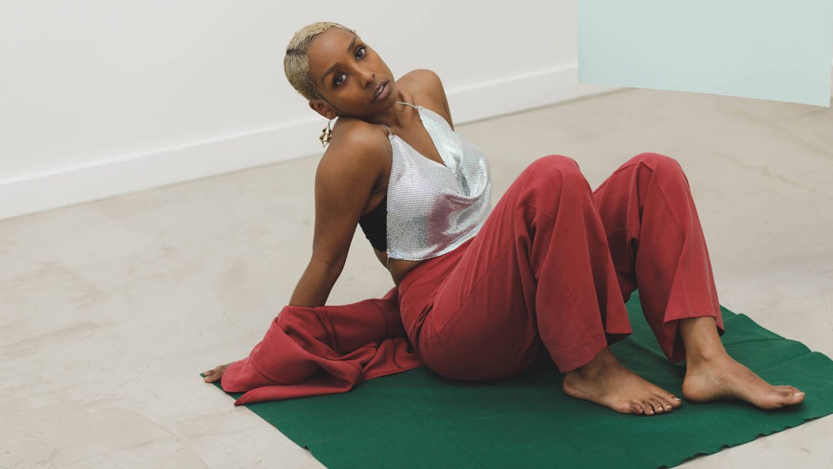 Person with blonde hair wearing a silver halter top and red pants sitting on a green mat, leaning back on hands, with bare feet.