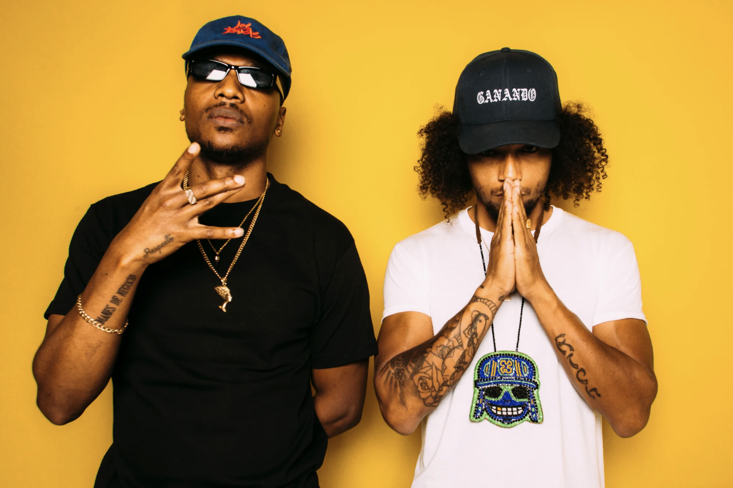 Two men posing in front of a yellow background, wearing hats and casual clothing. The man on the left is in a black shirt making a hand gesture, while the man on the right, wearing a white shirt, has his hands together in a prayer position. Both have tattoos and necklaces.