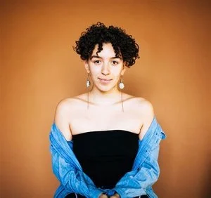 A person with short curly hair and dangling earrings wearing a strapless black top and a blue denim jacket draped over their shoulders, posing against a warm brown background.