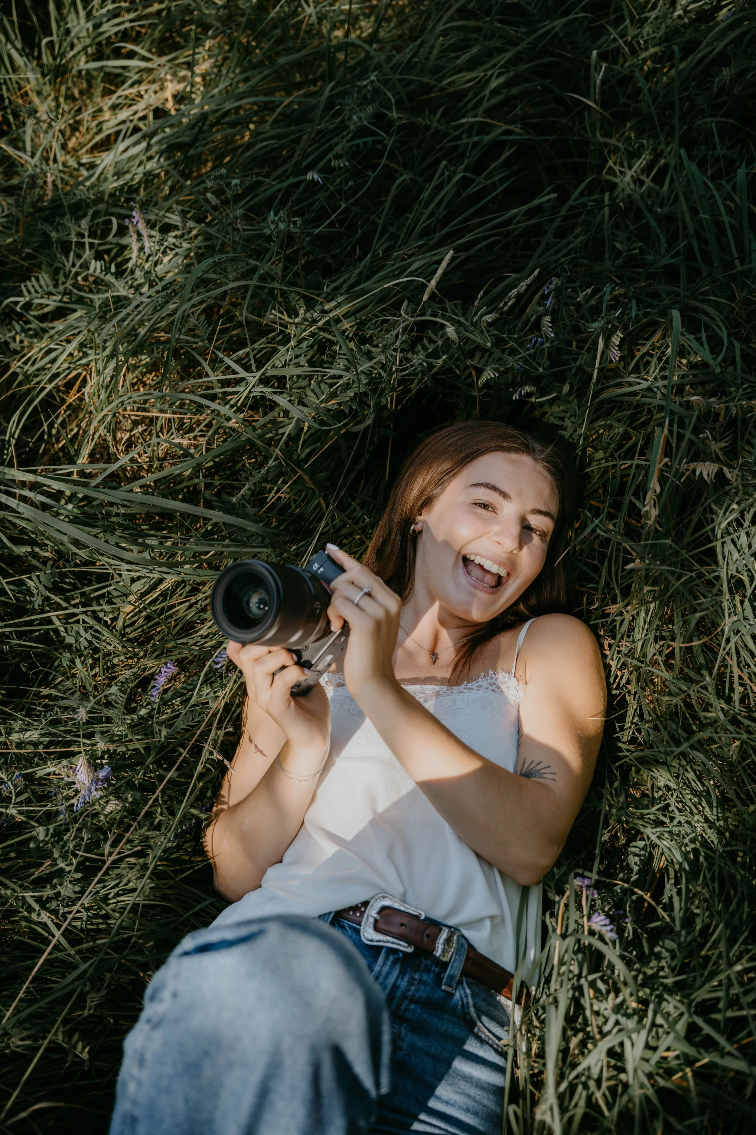 A woman with long brown hair lying in tall grass holding a camera and smiling.