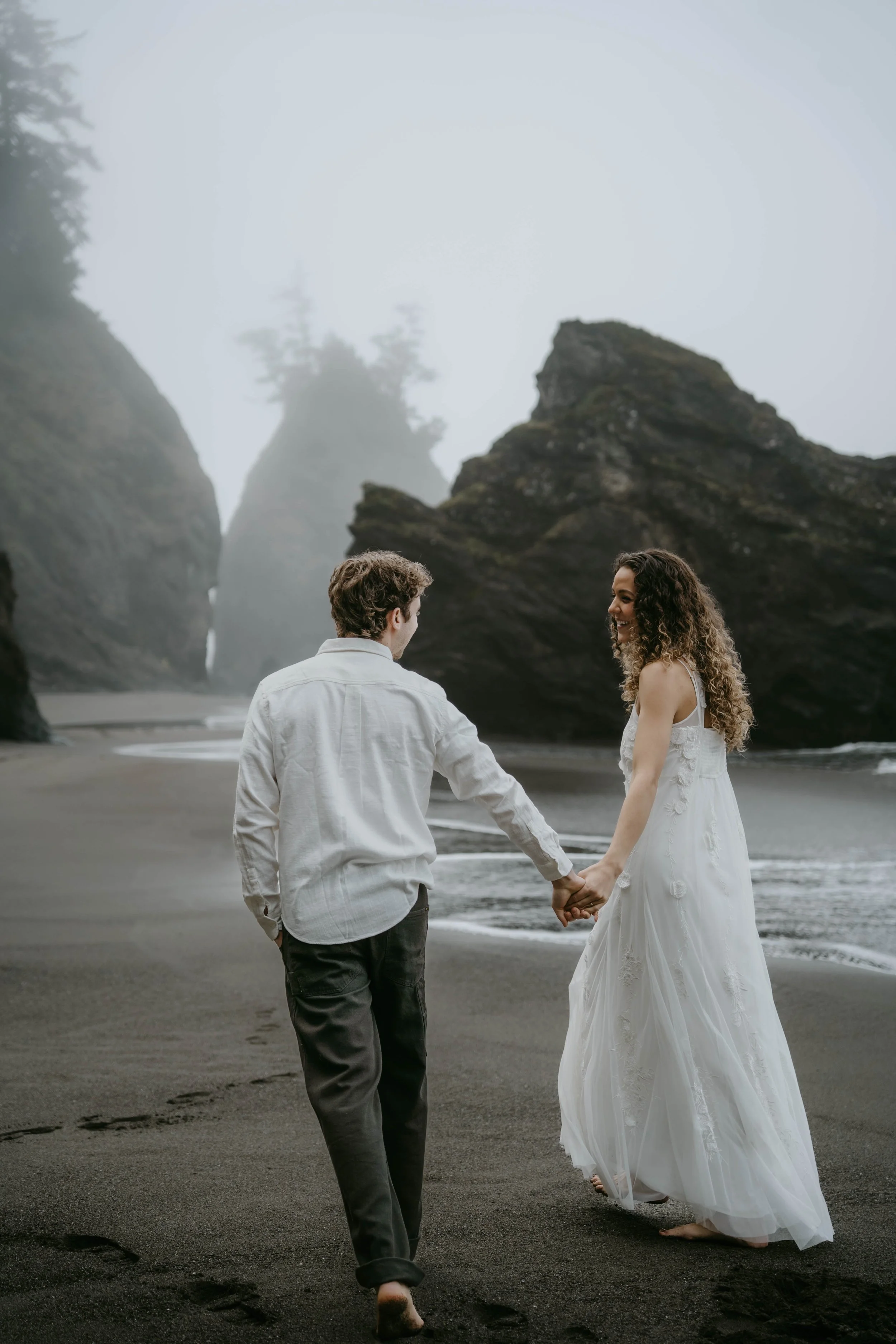 A couple holding hands walking on a black sand beach with large rock formations and misty skies in the background. The woman is wearing a white dress, and the man is dressed in a white shirt and dark pants.
