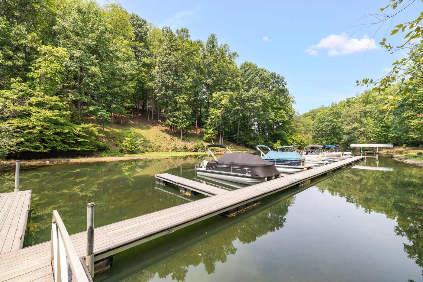 A private dock lined with covered pontoon boats extends into a calm green lake bordered by wooded hills under a blue sky.