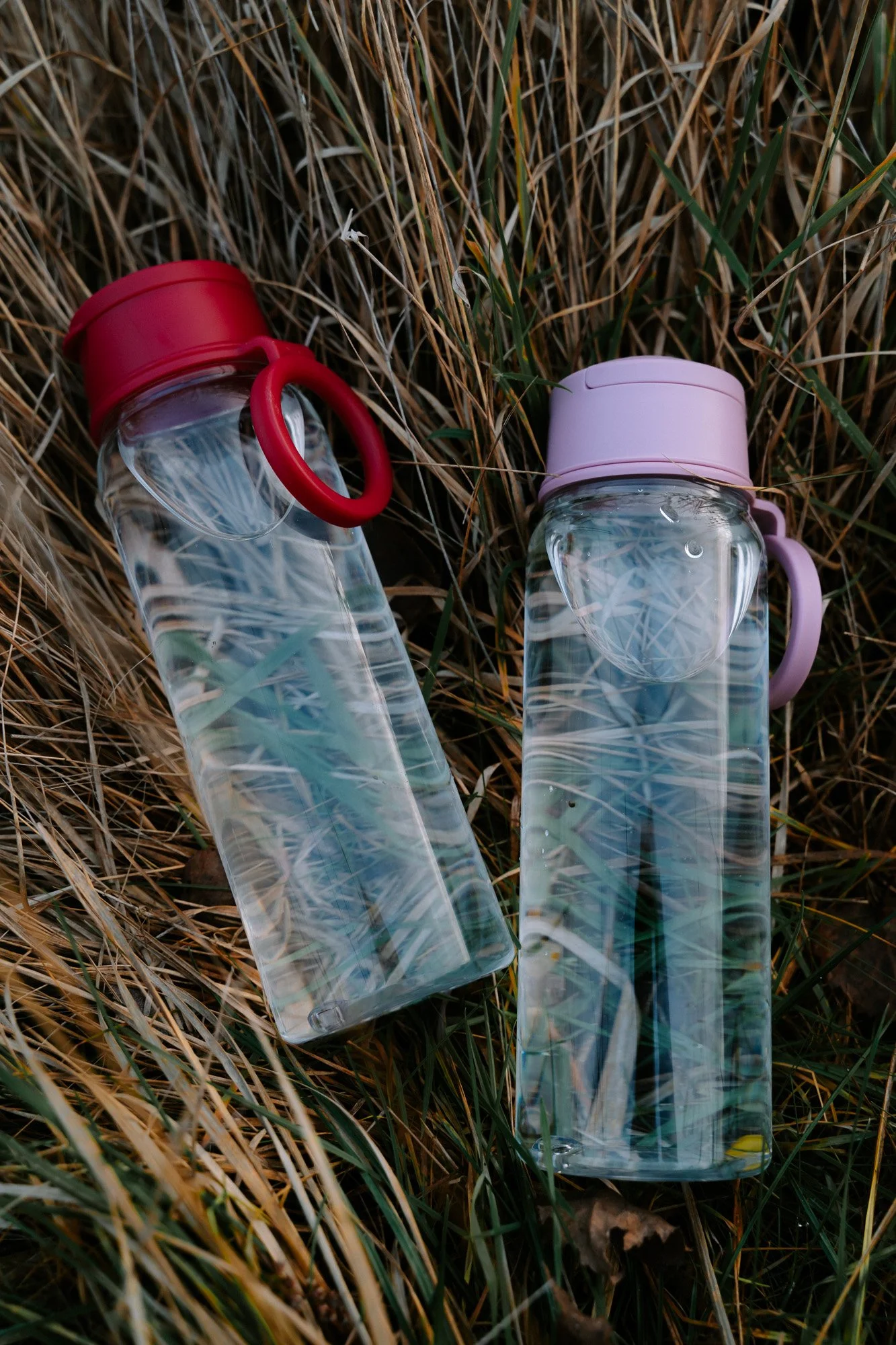 Twee doorzichtige waterflessen met gekleurde deksels en gemakkelijk los te draaien handvatten liggen in het gras.