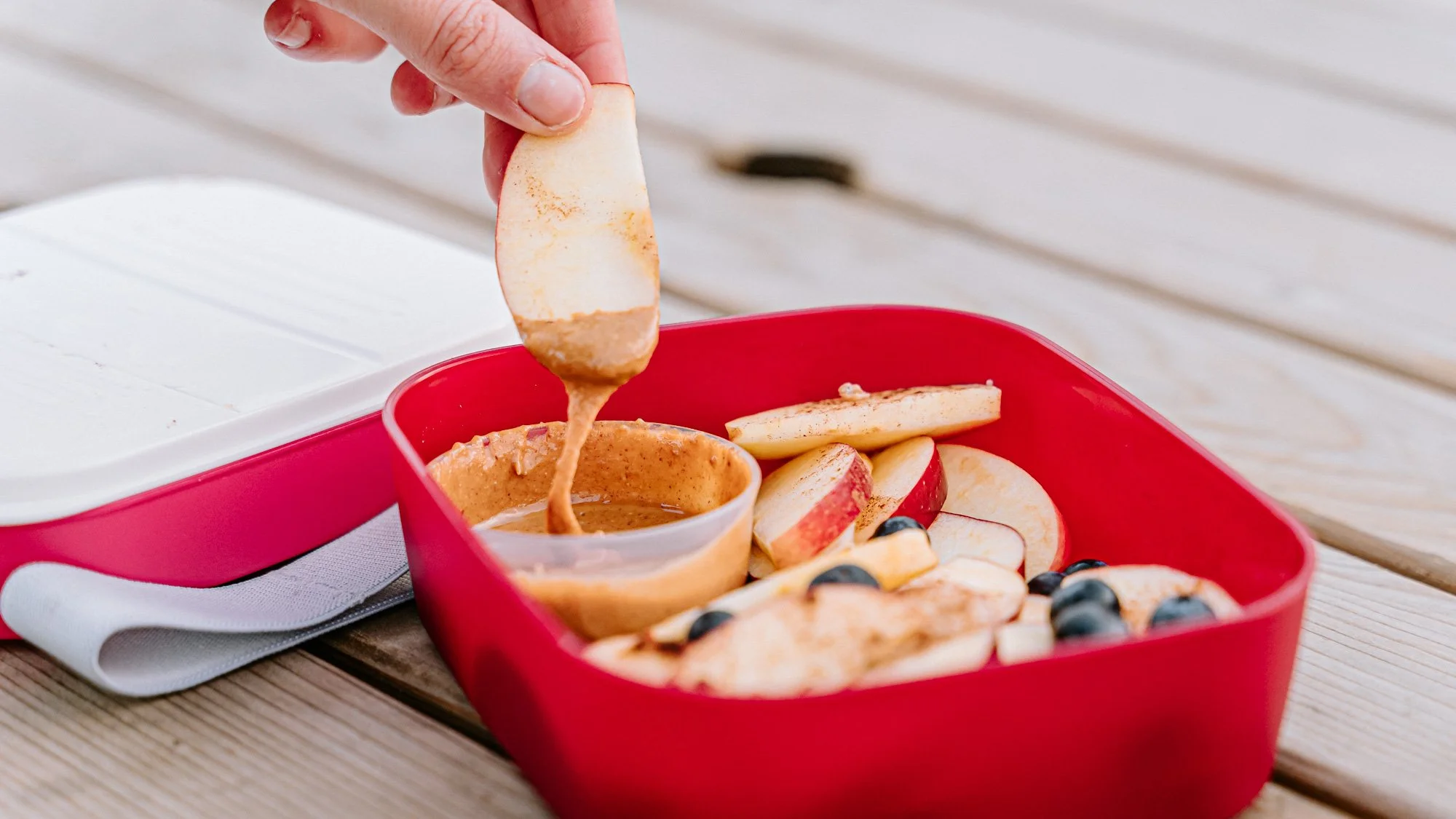 Een hand doopt een schijfje appel in pindakaas, die in een rode snackbox ligt samen met meer appelpartjes, bosbessen en een kommetje pindakaas.