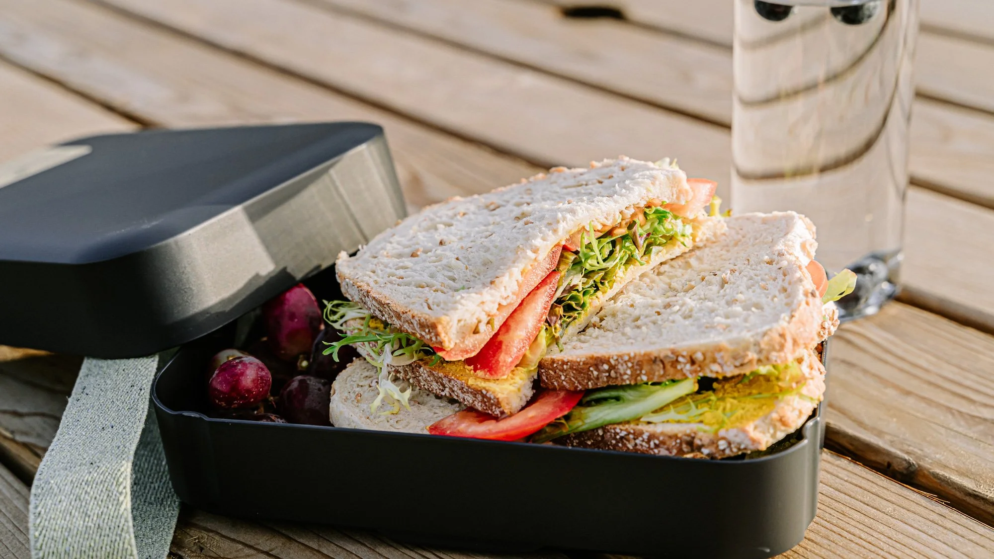 Twee sandwiches met vlees en salade, liggen in een zwarte brooddoos naast wat blauwe besjes en een glas water op een houten tafel.