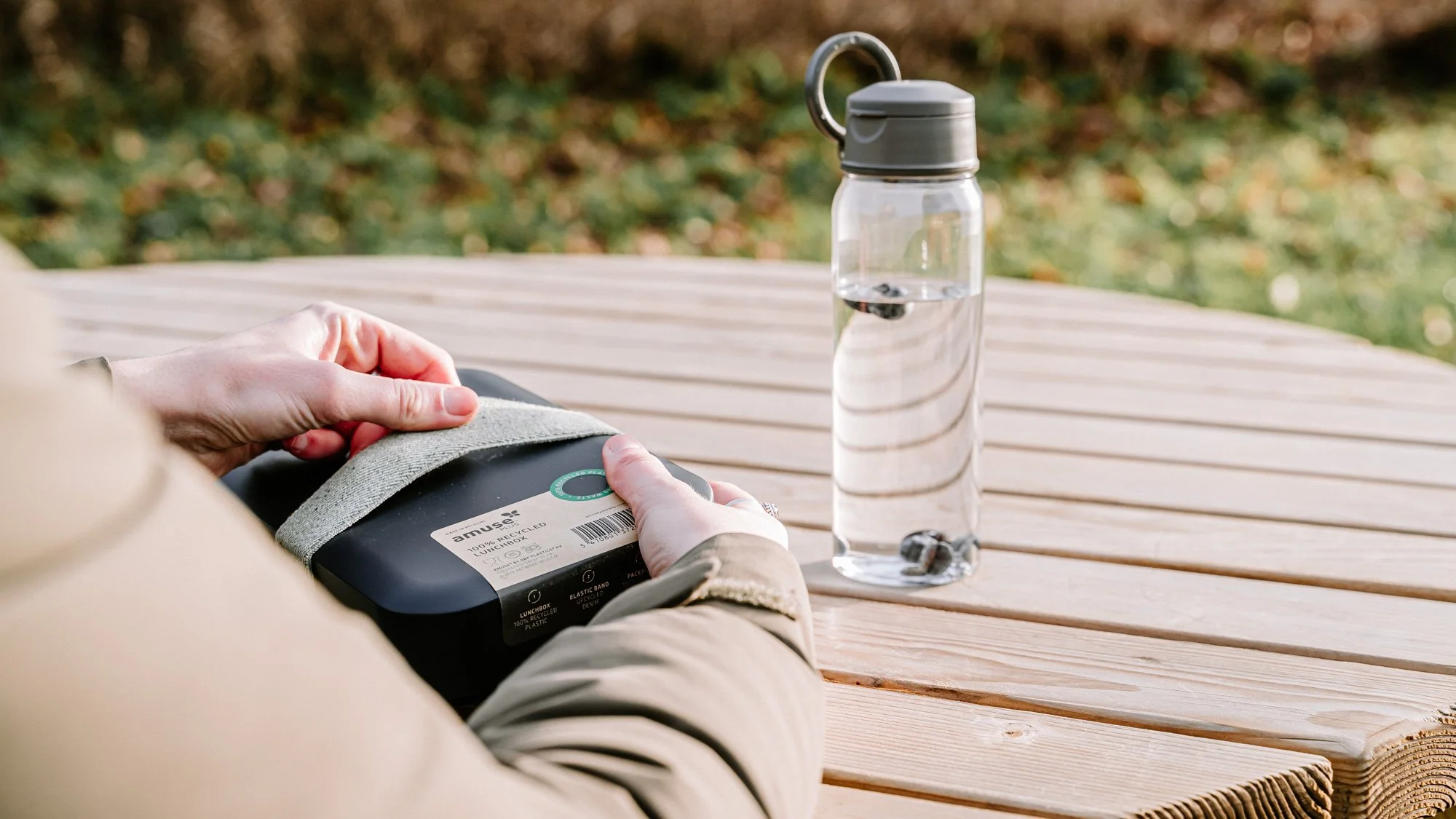 Persoon die een voedselpakket uitpakt op een houten tafel buiten, met een waterfles naast hen.