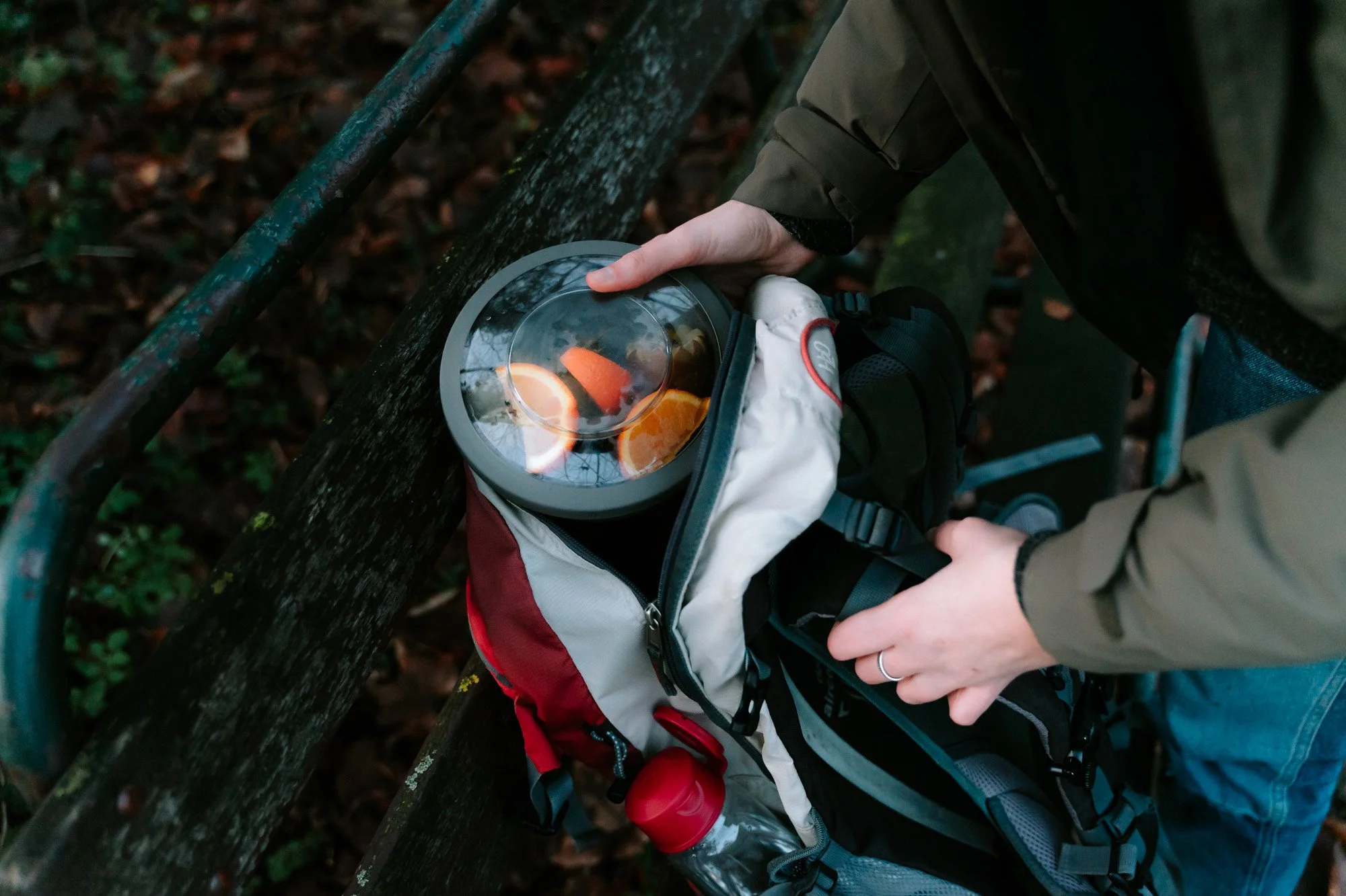 Persoon pakt een voedselcontainer uit een rugzak tijdens wandeltocht in bos, met een waterfles en een bankje op de achtergrond.