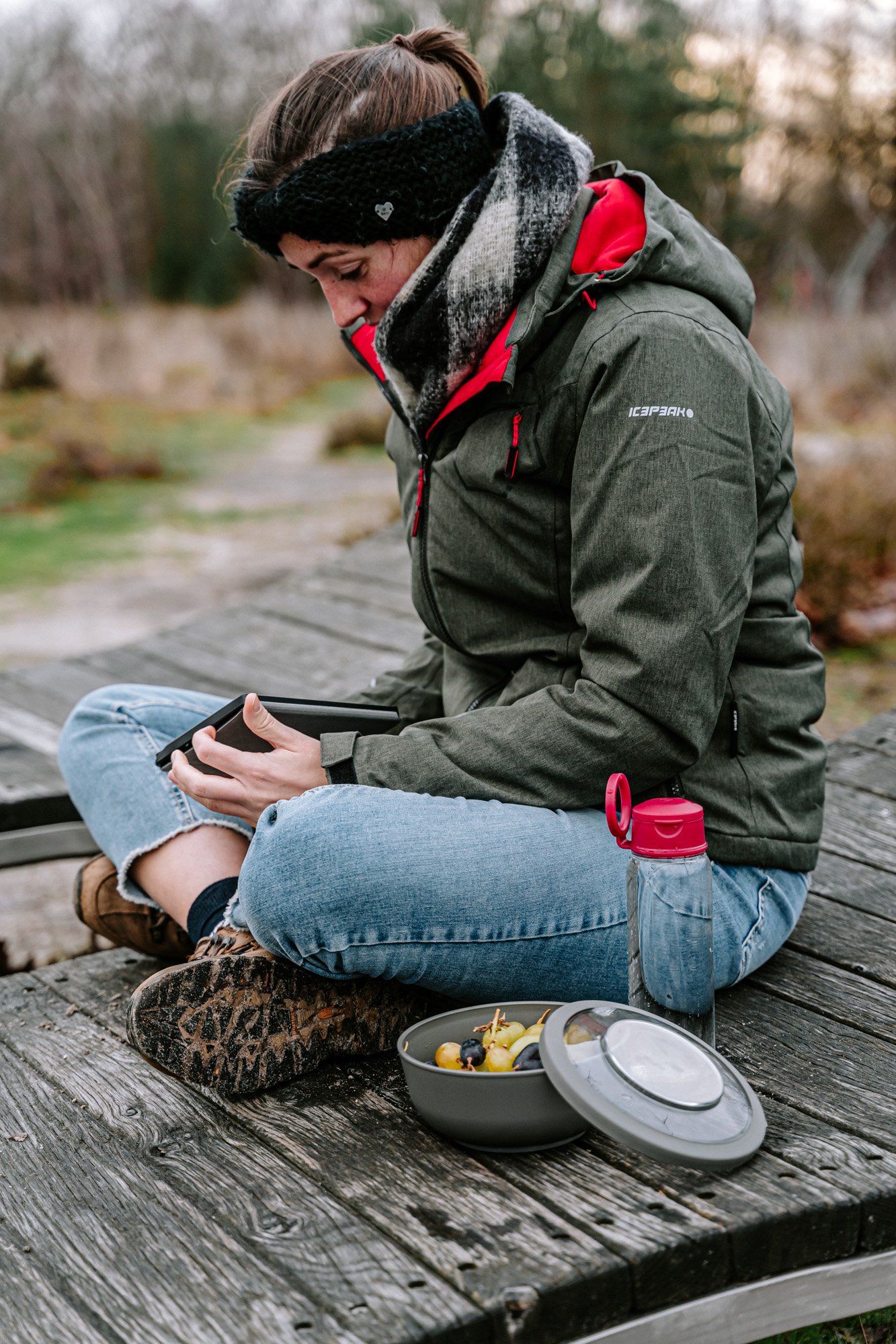 Vrouw zit op een houten bank in de natuur, kijkt op haar telefoon, met een bakje fruit en een waterfles naast haar.