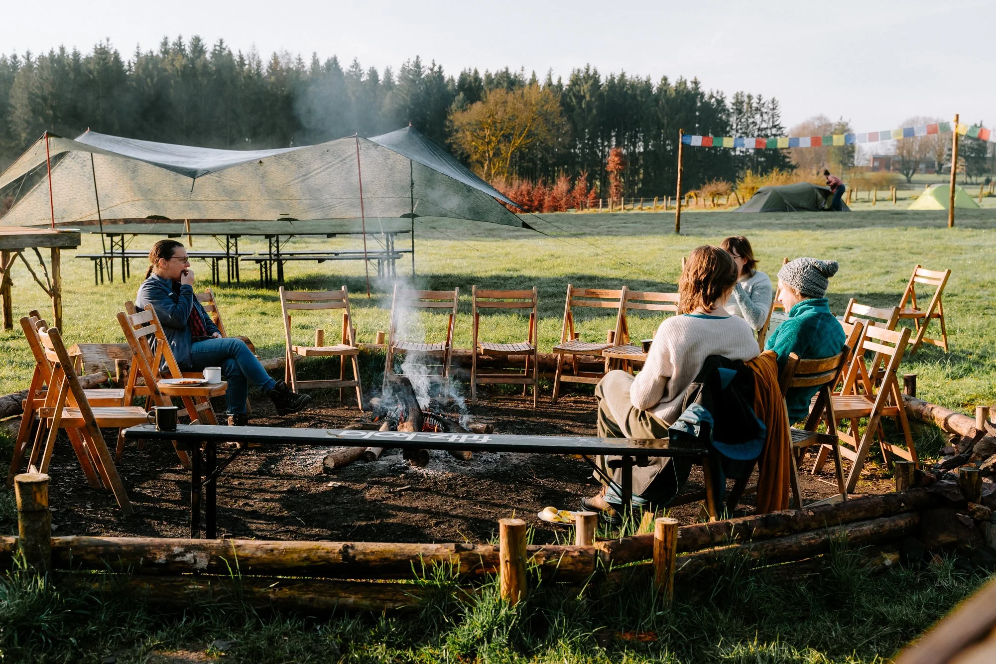 Vijf mensen zitten rond een kampvuur in een groene open ruimte met tenten en bomen op de achtergrond, genietend van een gezellige bijeenkomst.