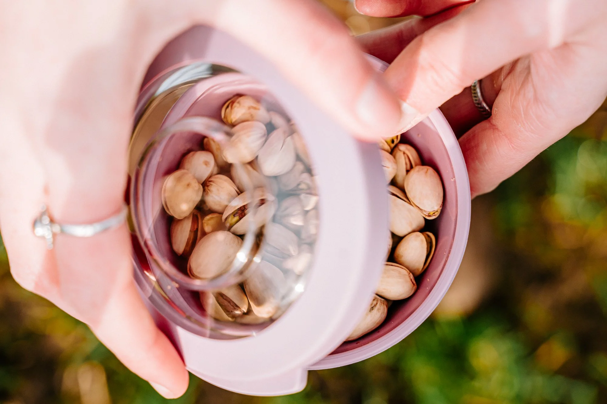 Close-up van een hand die een roze bak met pistachenoten vasthoudt, met een kleine glazen potje met water en pistachenoten glijdend naar beneden, achtergrond met gras en bladeren.