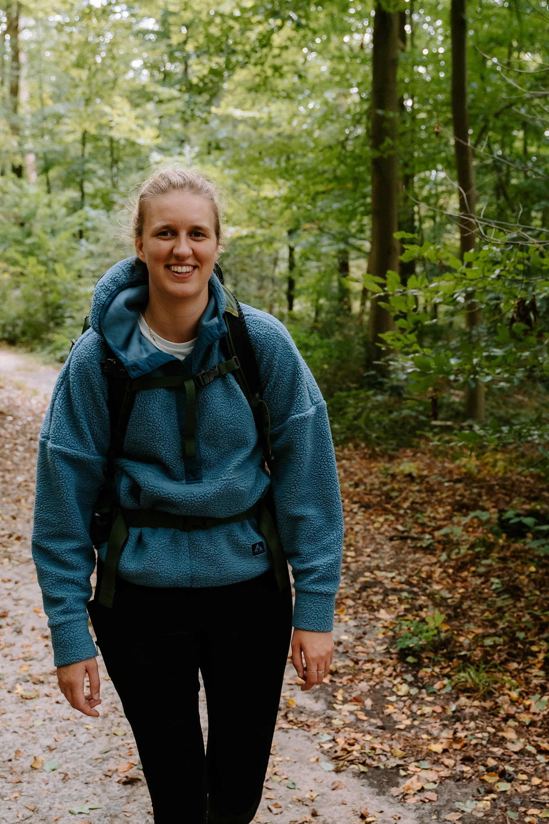 Vrouw met een blauwe gewatteerde jas en rugzak wandelt door een bos met groene bladeren en een gemarkeerd pad.