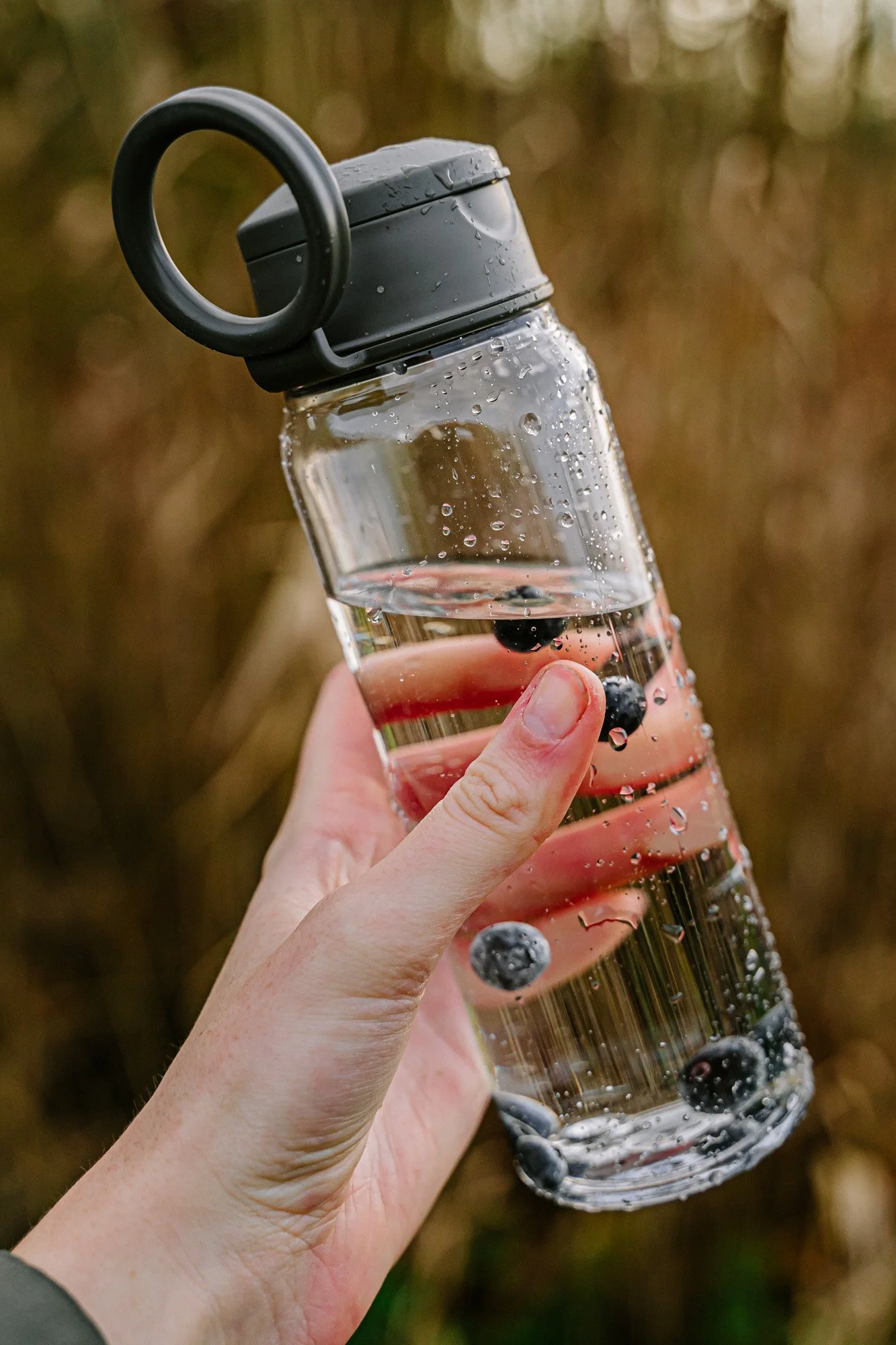 Persoon houdt een doorzichtige waterfles met een grijze drinkdop en een ring vast, gevuld met water en bosbessen, buiten met een bokeh van herfstbladeren op de achtergrond.