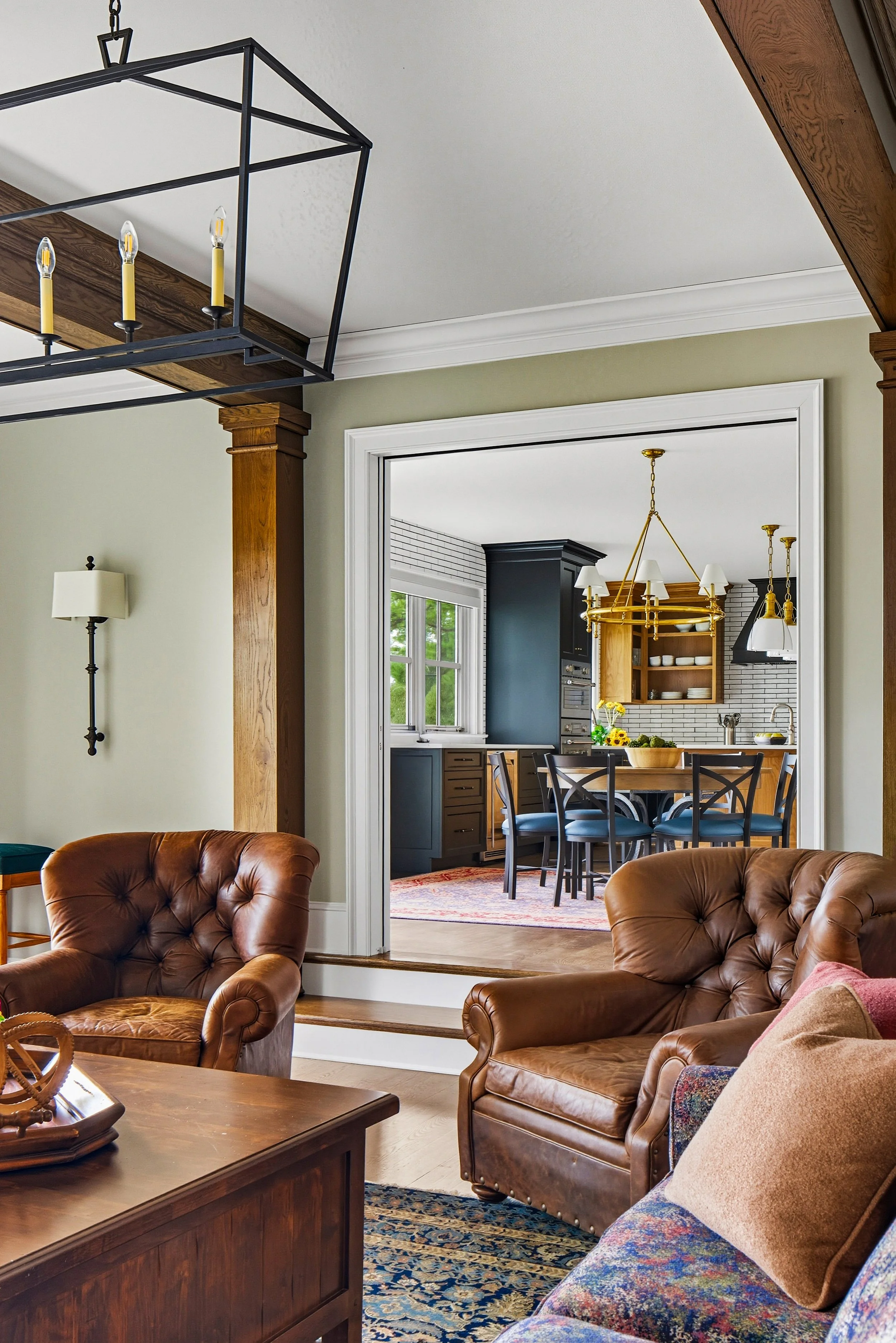 Living room with brown leather armchairs, a wooden coffee table, and a view into a dining area with a round wooden table, black chairs, and a chandelier, kitchen, window, and black cabinets in the background.