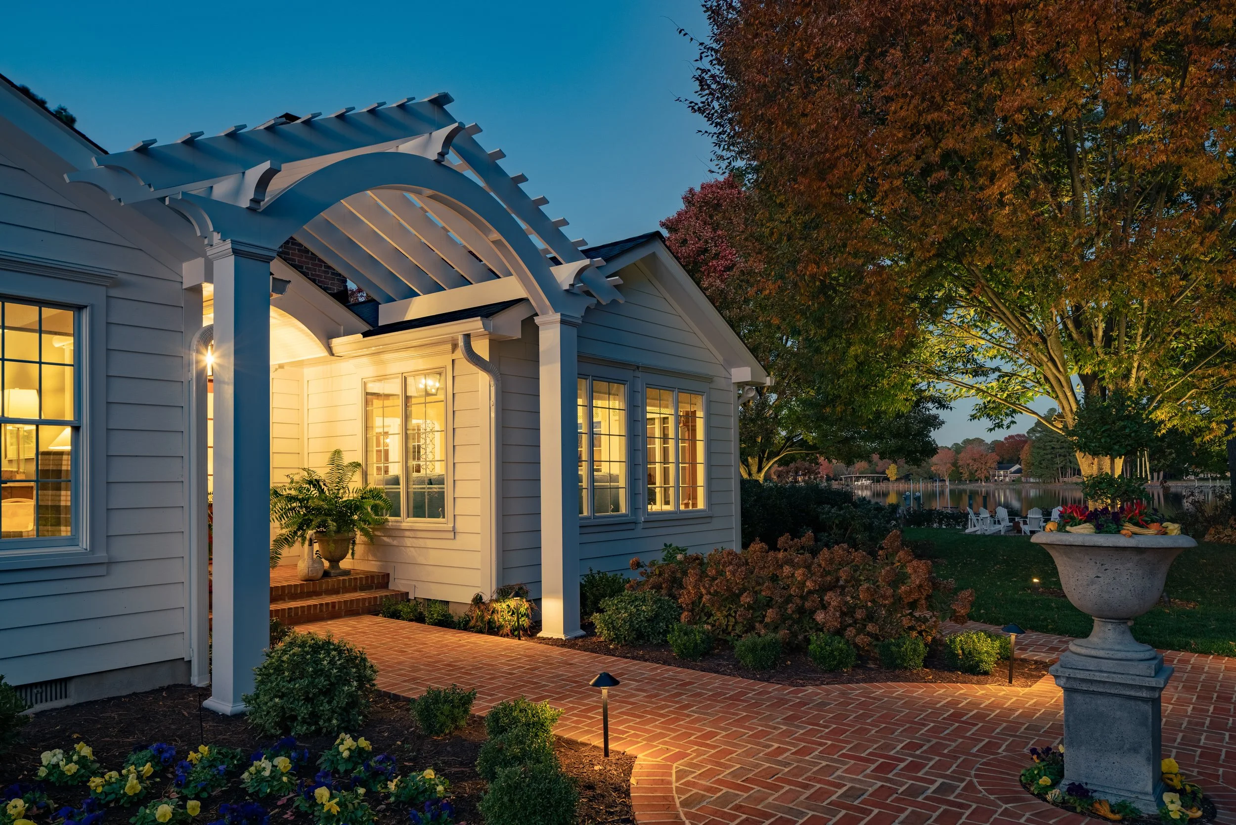 Exterior view of a house with illuminated windows, a brick pathway, potted plants, trees, and a lakeside in the evening.