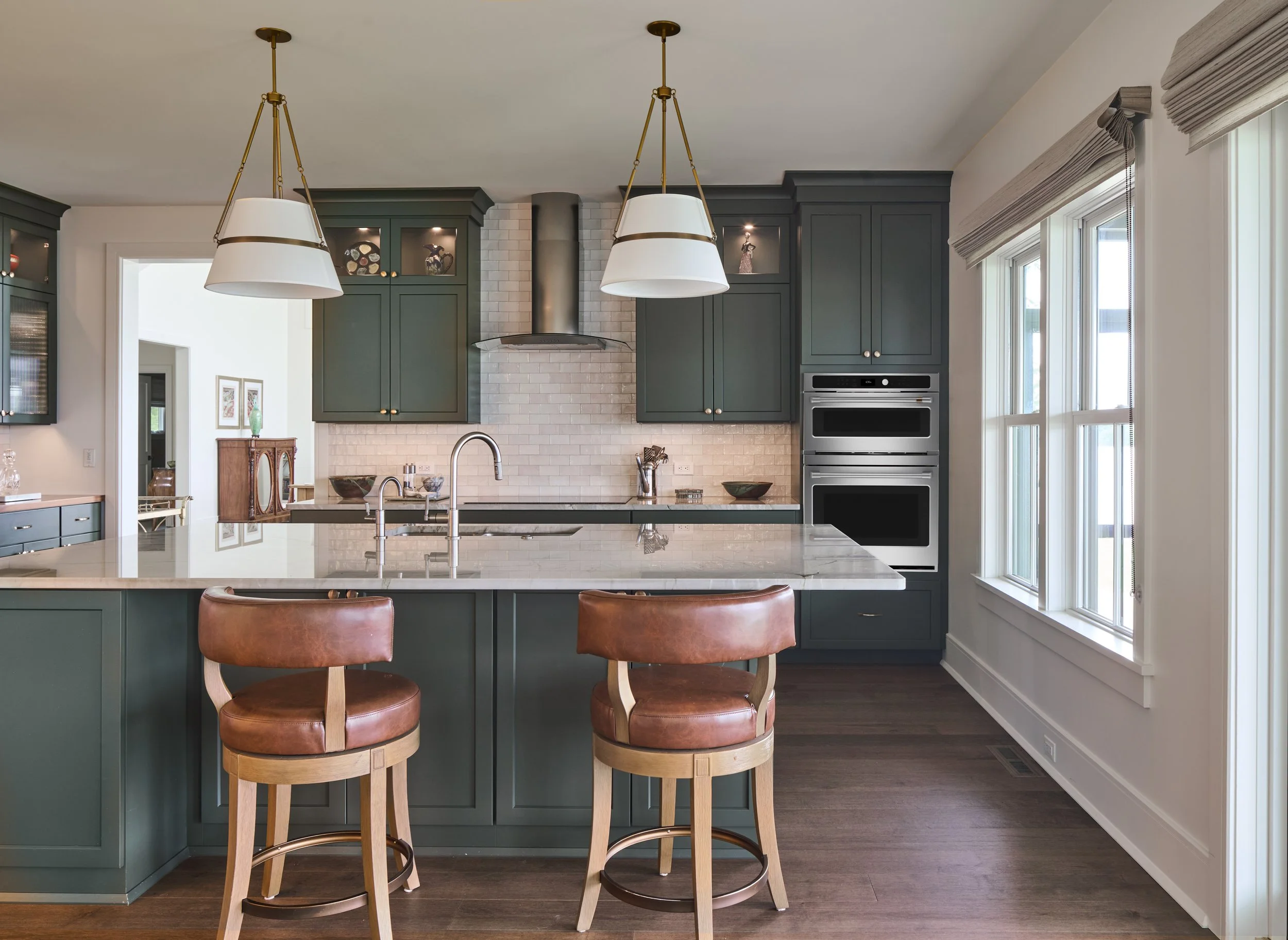 Modern kitchen with green cabinetry, white marble island, brown leather barstools, large windows, and stainless steel appliances.