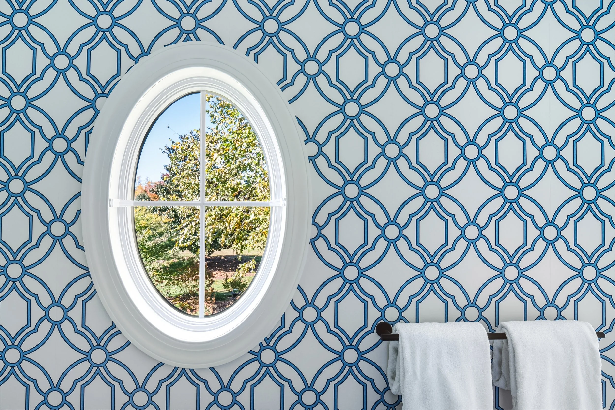 Bathroom wall with blue geometric patterned wallpaper, white oval window with cross trim showing outdoor trees and blue sky, and white towels hanging on a black towel rack.