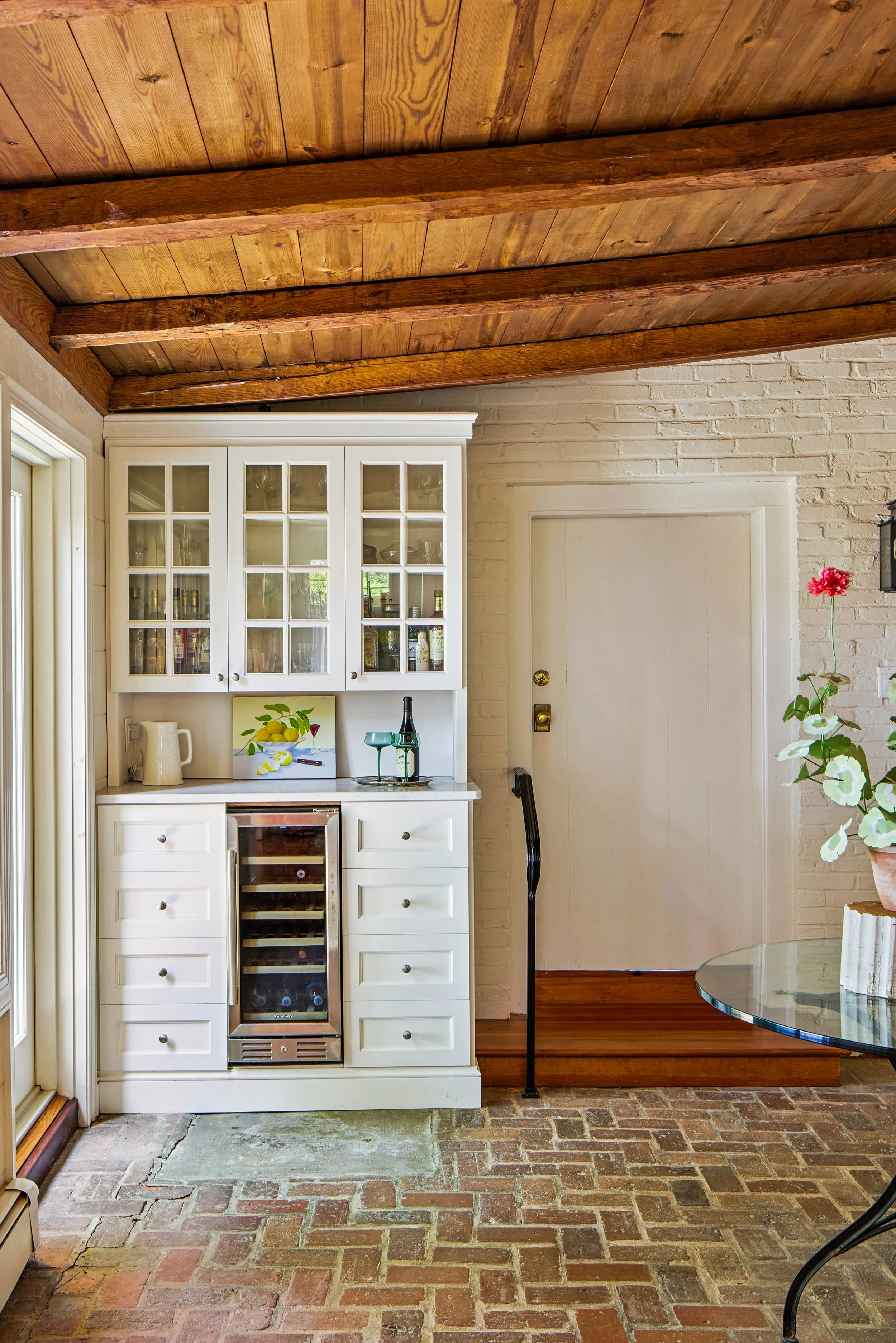 Interior of a kitchen featuring a white cabinet with glass doors, wine cooler, brick flooring, wooden ceiling beams, and a glass table with a potted flower.