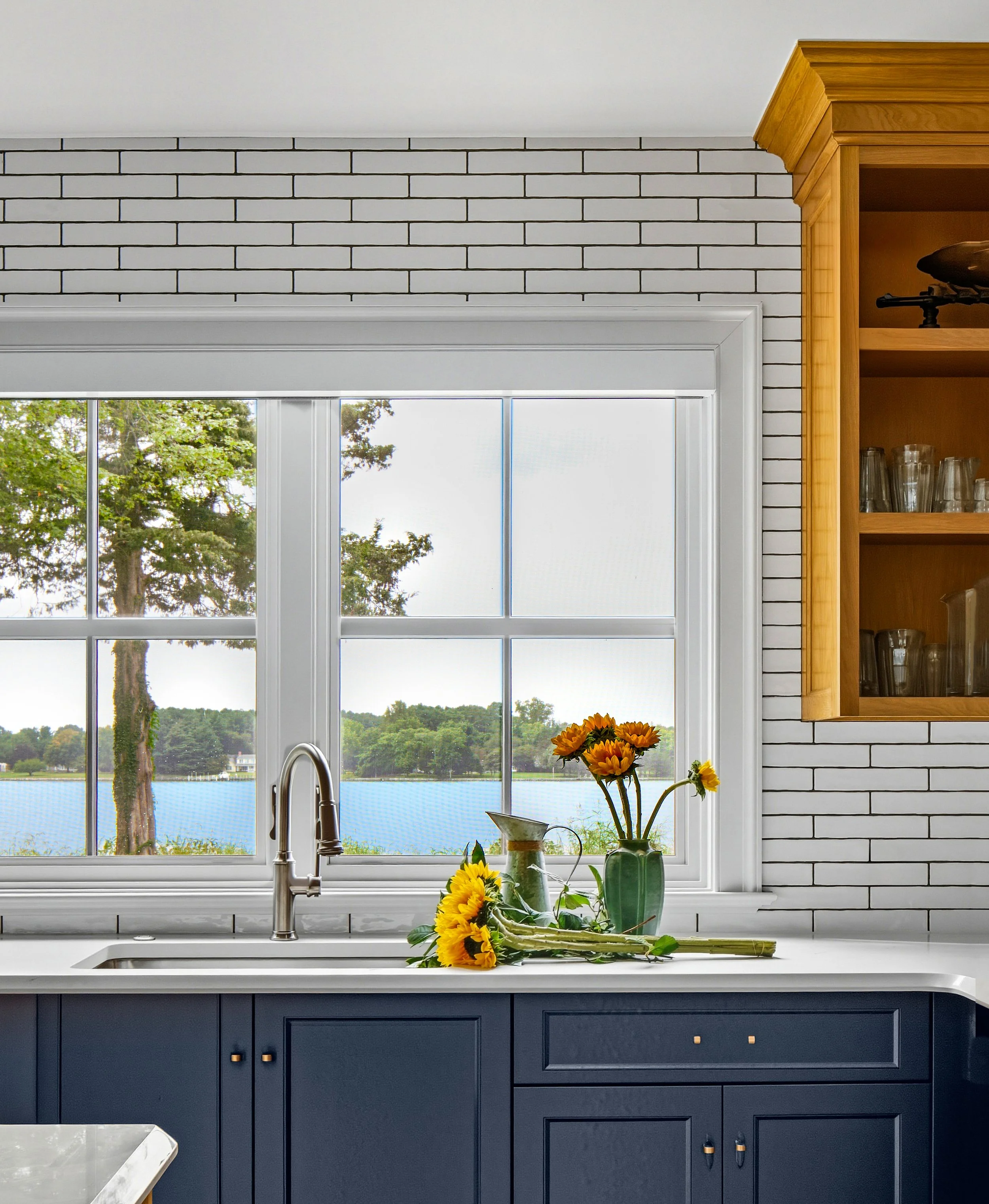 Kitchen with navy blue cabinets, white countertop, a window overlooking a lake with trees, and flowers in vases on the counter.