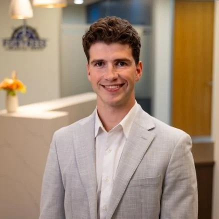 A young man in a light gray suit and white shirt smiling in an indoor office setting.