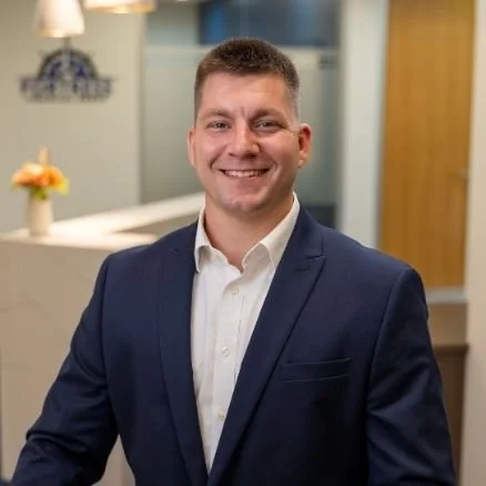 A man in a dark blue suit with a white shirt, smiling in an indoor setting with a modern background, possibly an office or reception area.