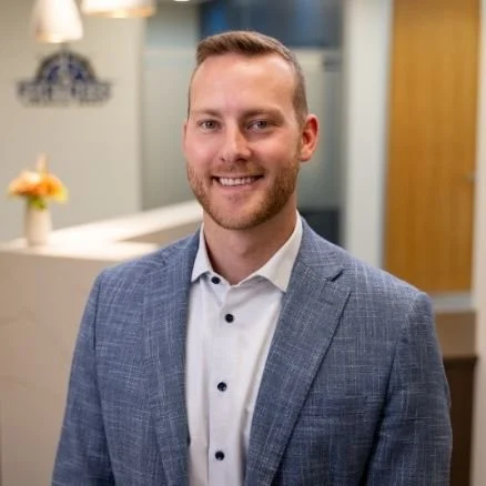 A man in a light blue suit jacket and white shirt smiling in an office environment.
