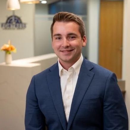 A young man in a blue suit and white shirt smiling in an office lobby, with a reception desk and a flower arrangement in the background.