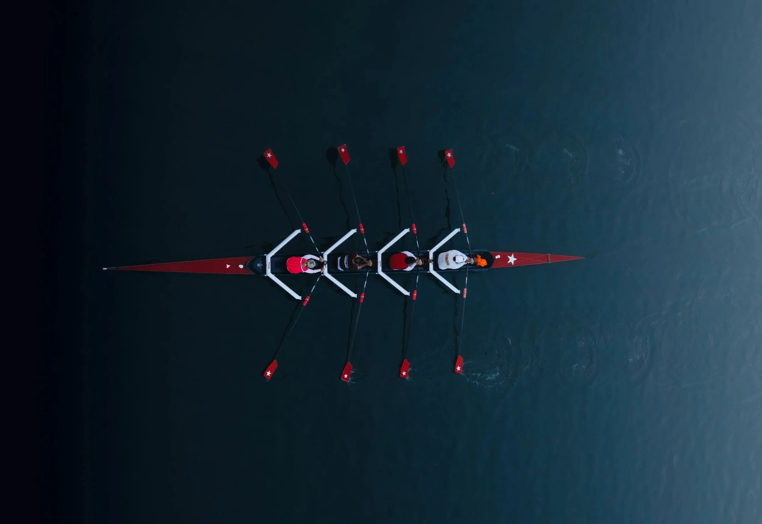 An overhead view of a rowing team in a red and white boat with four rowers and a coxswain, on dark water.