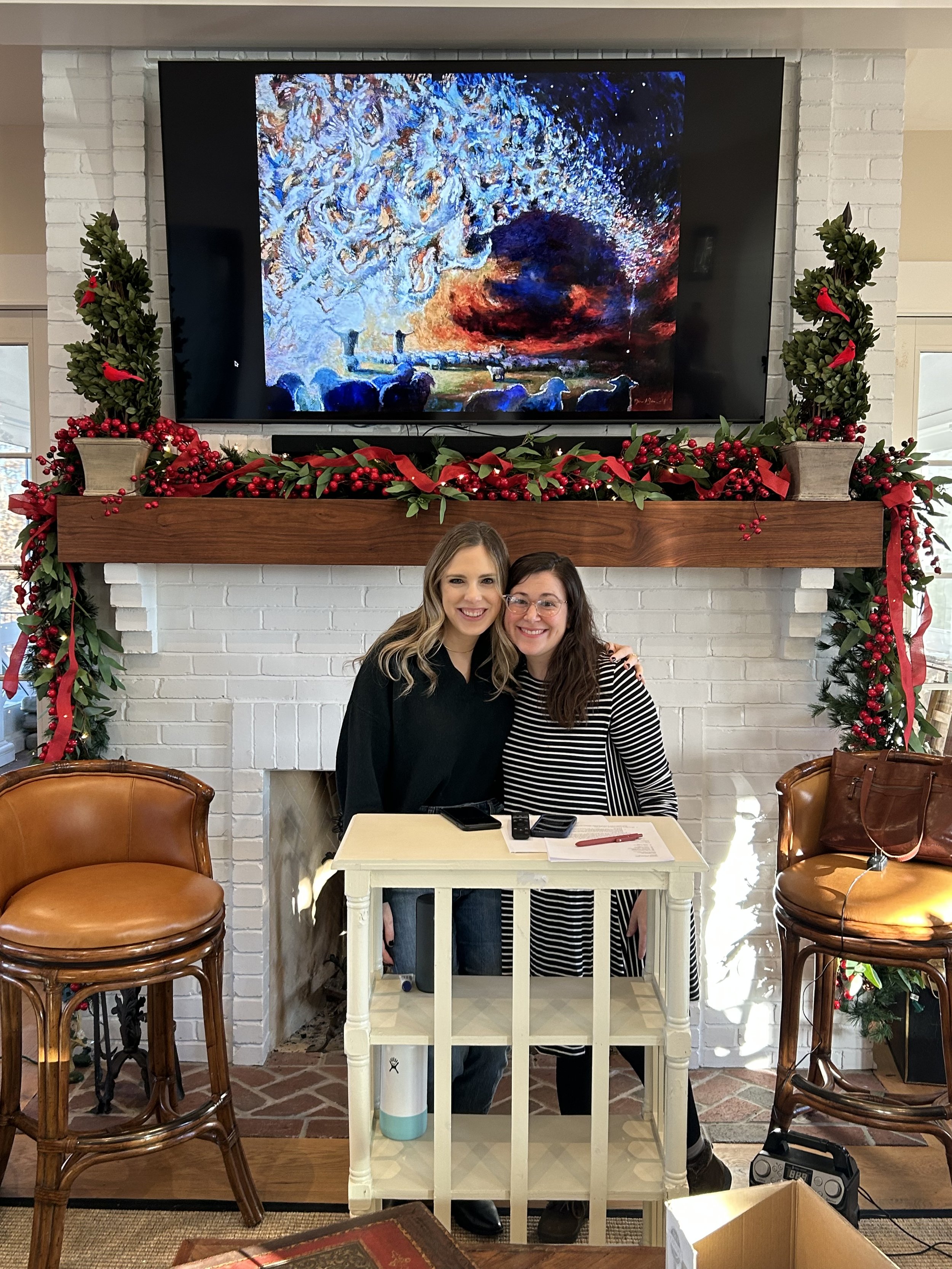 Two people smiling and posing for a photo in front of a decorated fireplace with holiday garlands and a flat-screen TV displaying a colorful artwork. There are two chairs, a table with papers and phones, and a brown bag in the scene.