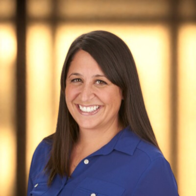 A woman with long dark hair, smiling, wearing a blue shirt, standing in front of an out-of-focus lit background.